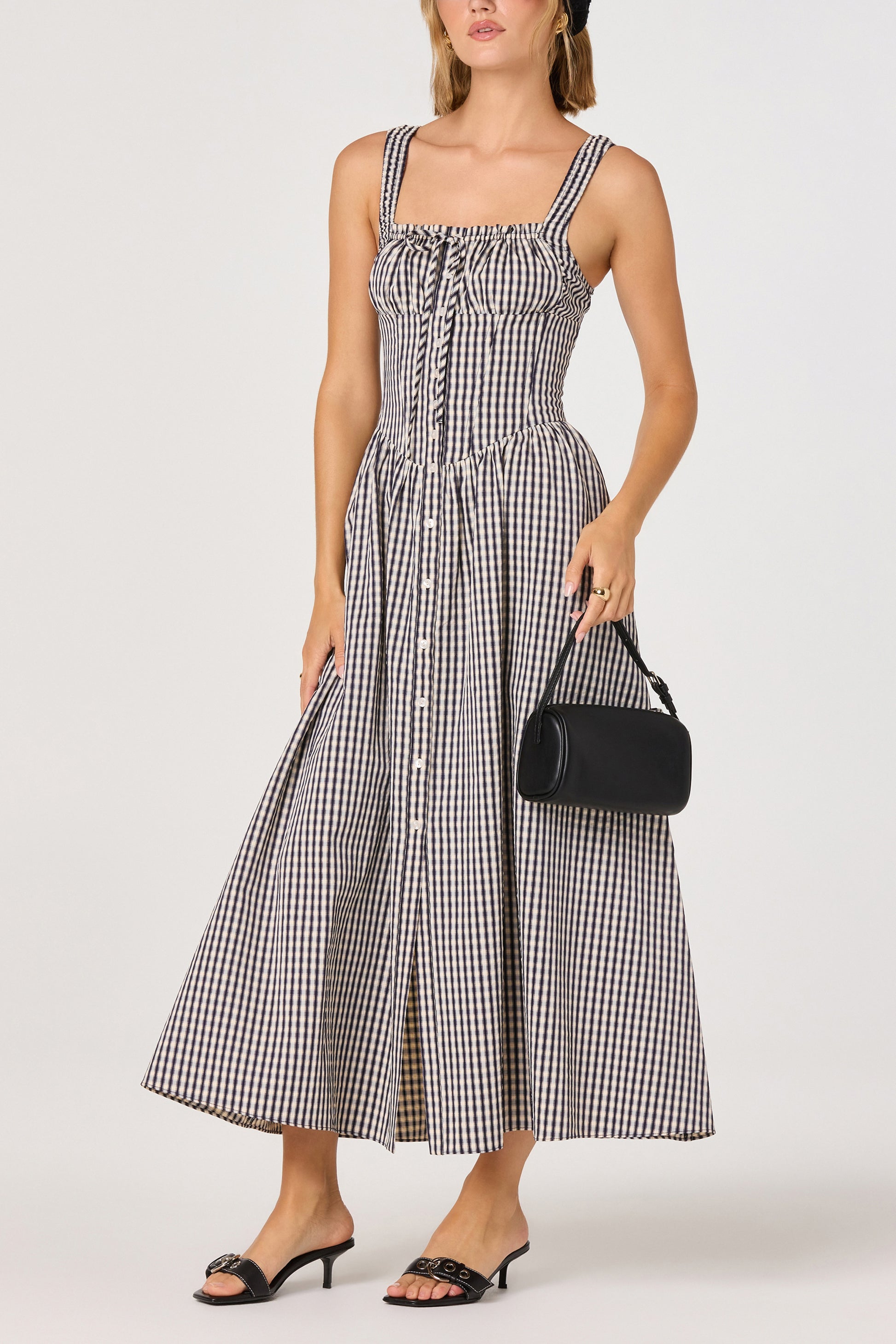 A woman wearing the ASTR Temperance Maxi Dress—a black and white gingham woven maxi dress with smocked tank straps and a fitted milkmaid bust—paired with a small black handbag and black heeled sandals.