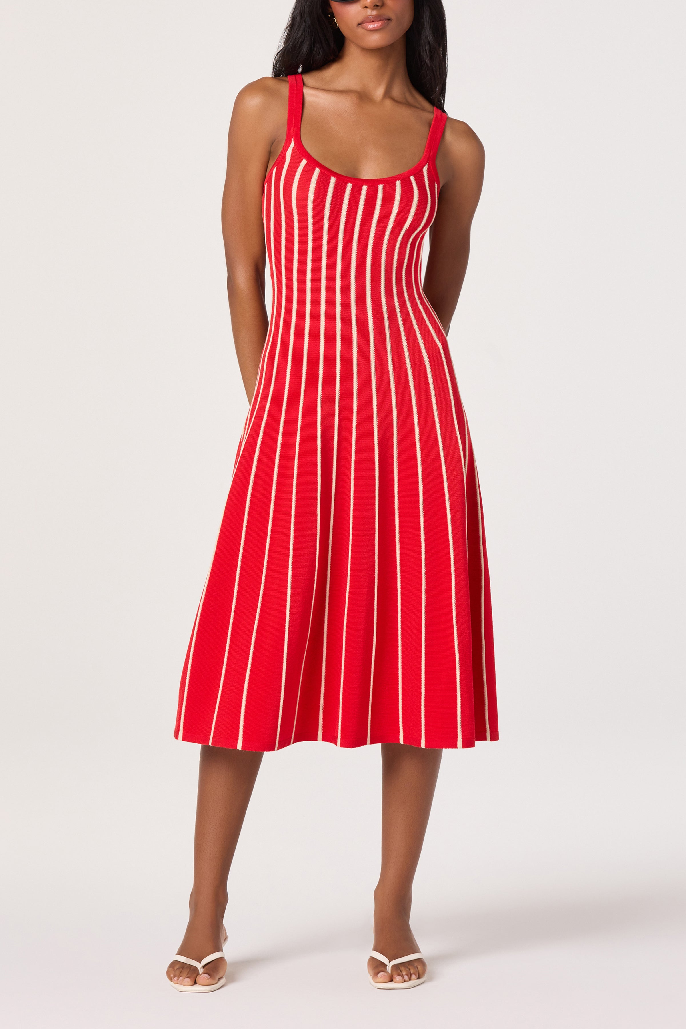 A woman wears the ASTR Irecia Sweater Midi Dress—a sleeveless, slim fit red dress with vertical white stripes and a scoop neckline—paired with white open-toe sandals, against a plain light background.