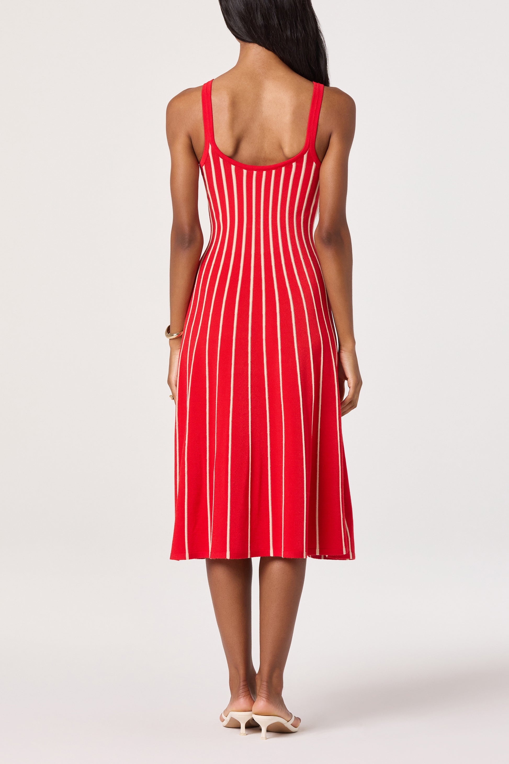 A woman with long dark hair, facing away, wears the ASTR Irecia Sweater Midi Dress—a sleeveless red ribbed knit with vertical white stripes and a scoop back—paired with white heeled sandals on a light background.