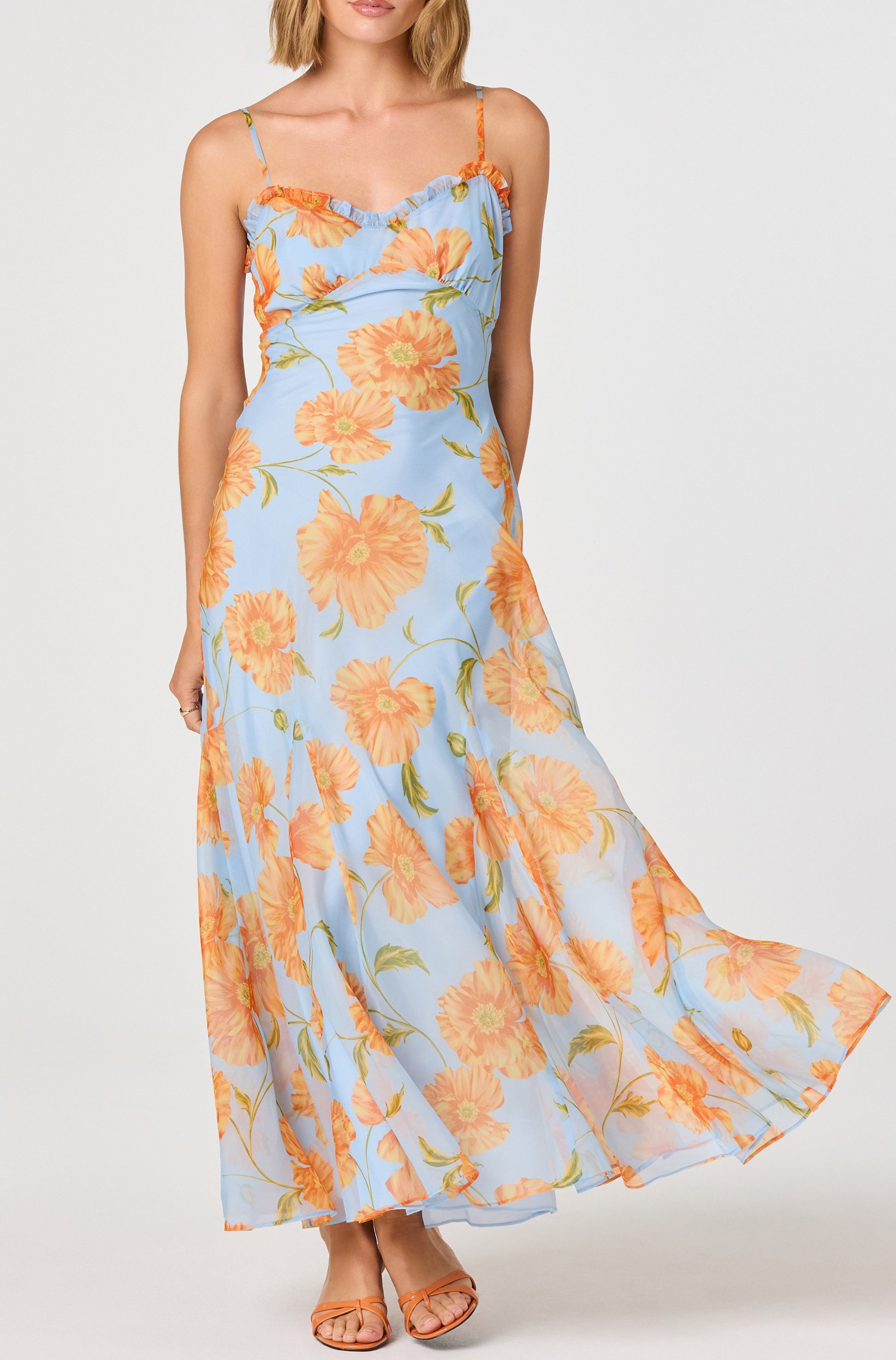 A woman models the ASTR Maeryn Maxi Dress, showcasing its orange floral print, sweetheart neckline, spaghetti straps, ruffled details, and flowing godet skirt. She stands against a white background in matching flat sandals.