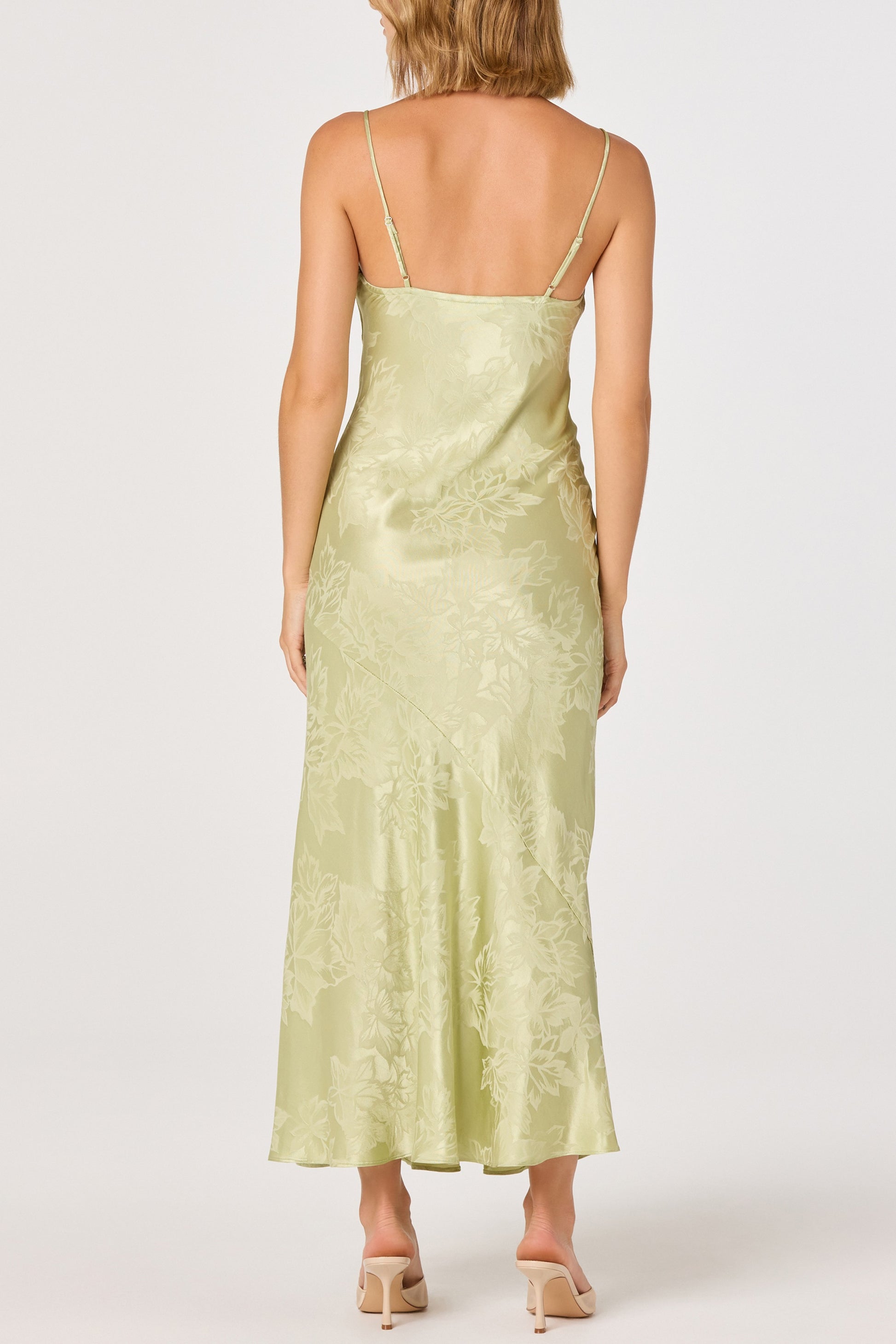 A woman with shoulder-length light brown hair wears the ASTR Tyla Midi Dress, a light green, bias-cut floral dress with thin straps and an open back, paired with beige heeled sandals.