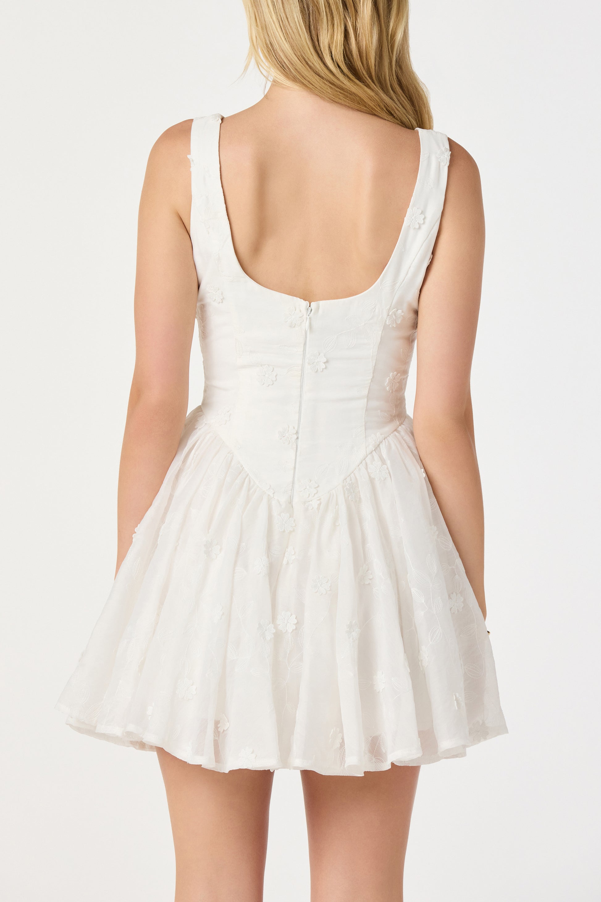 A woman with long blonde hair, seen from behind, wears the ASTR Reina Mini Dress—a sleeveless woven mini with a fitted bodice, flared above-the-knee skirt, subtle floral detail, scoop back, and zipper closure.