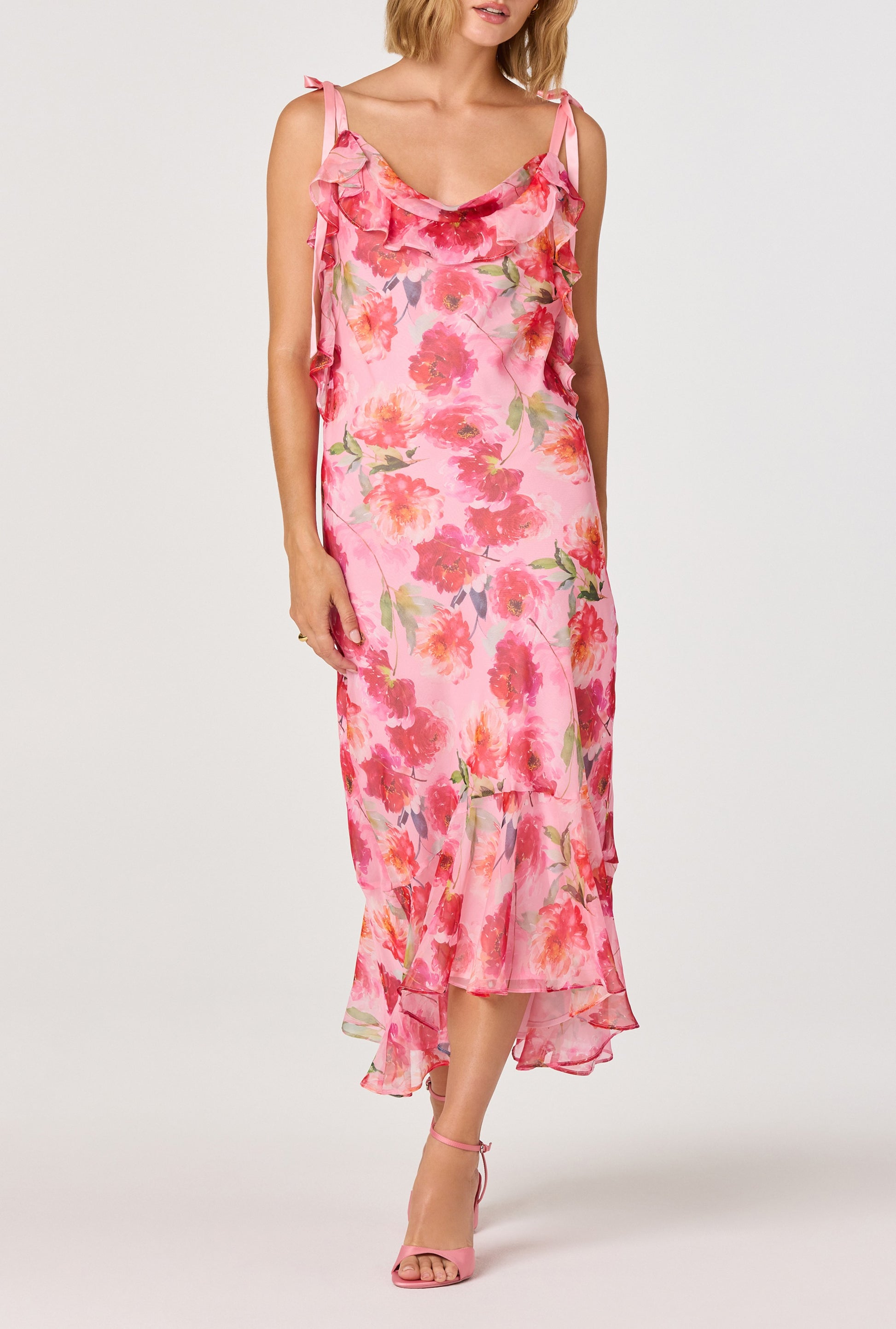 A woman wearing the ASTR Nissa Maxi Dress in pink floral with cascading ruffle trim and matching pink heels stands against a plain light background. Her face is partially out of frame.