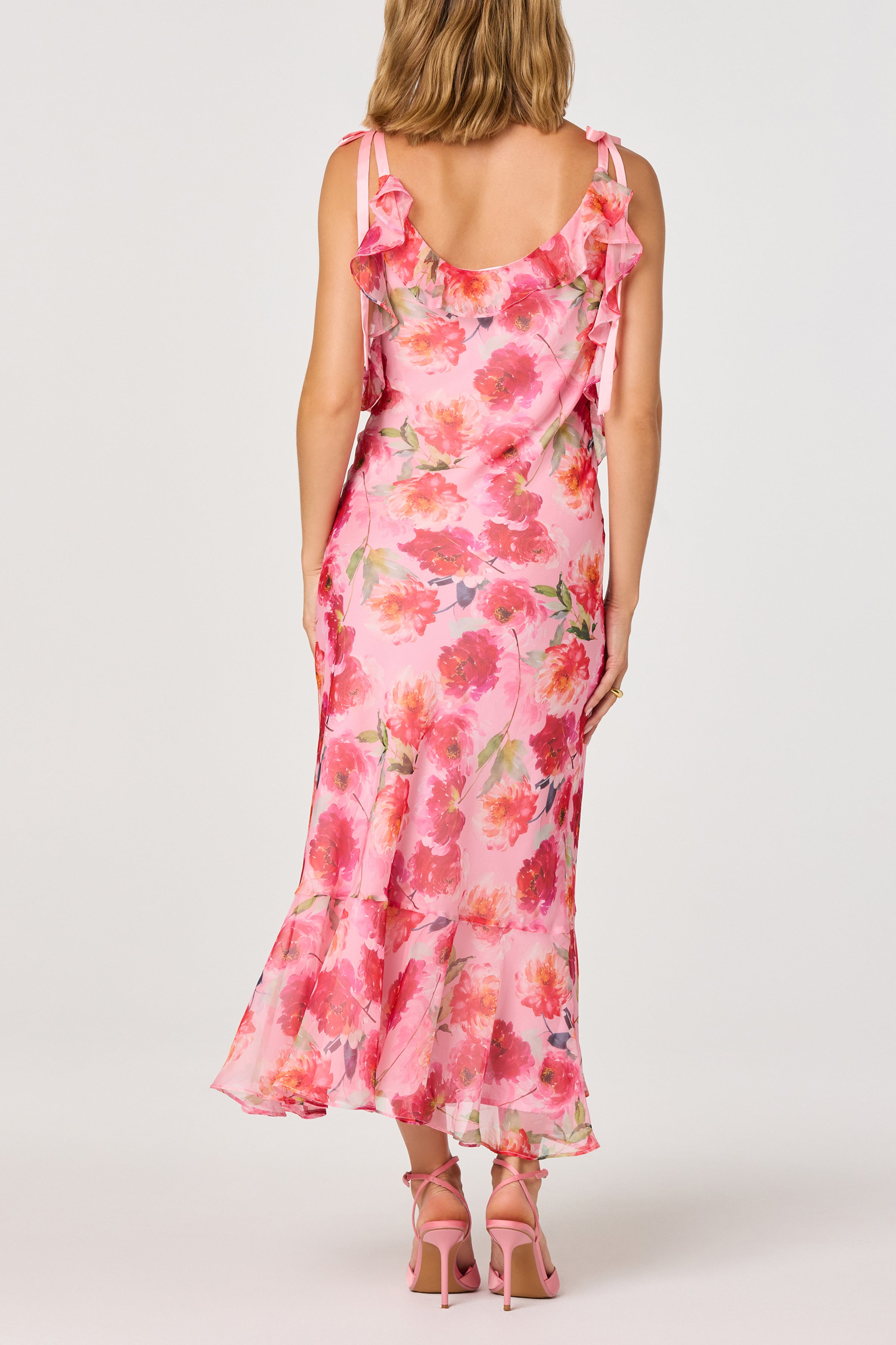 A woman stands facing away from the camera, wearing the ASTR Nissa Maxi Dress—a sleeveless pink floral dress with cascading ruffle trim and a flared hem—paired with pink high-heeled sandals.