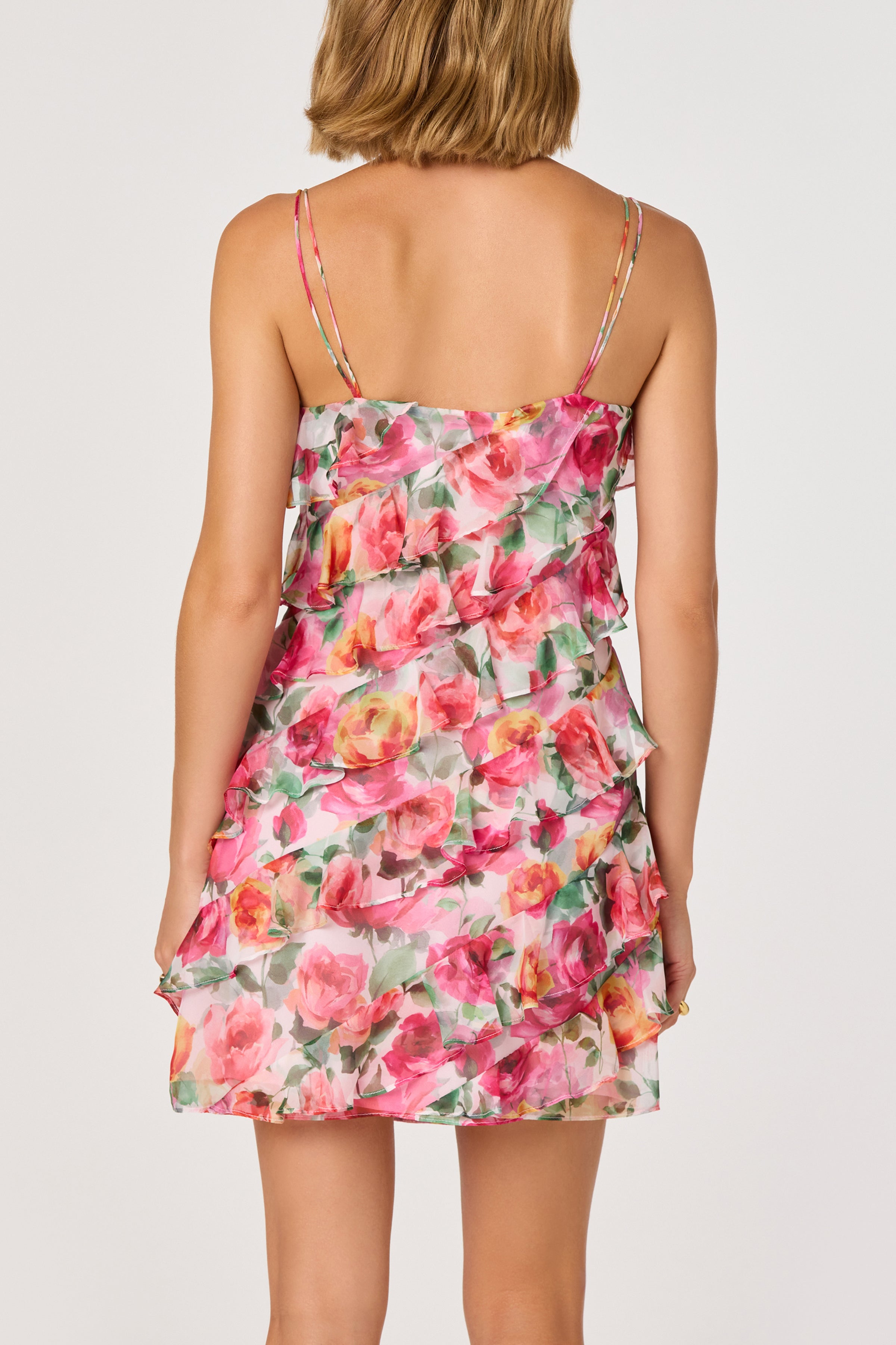 A woman with short blonde hair is seen from the back, wearing the ASTR Blanca Mini Dress—a sleeveless pink and green floral chiffon mini with thin straps, layered asymmetrical ruffles, and an above-the-knee hemline.