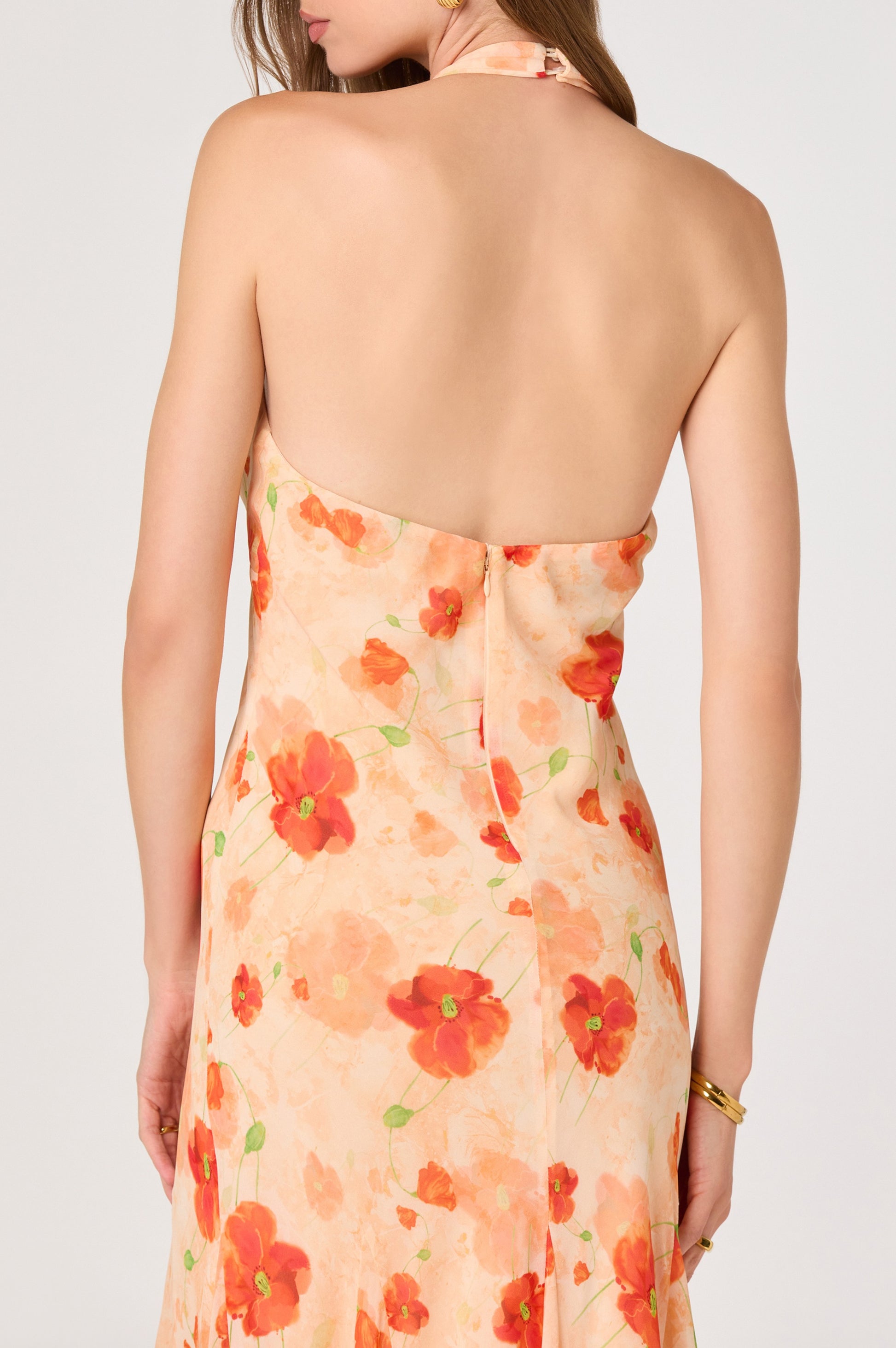 A woman stands with her back to the camera in the Yasmeen Maxi Dress by ASTR, featuring pleated straps, a backless halter design, and a peach and red floral chiffon fabric.
