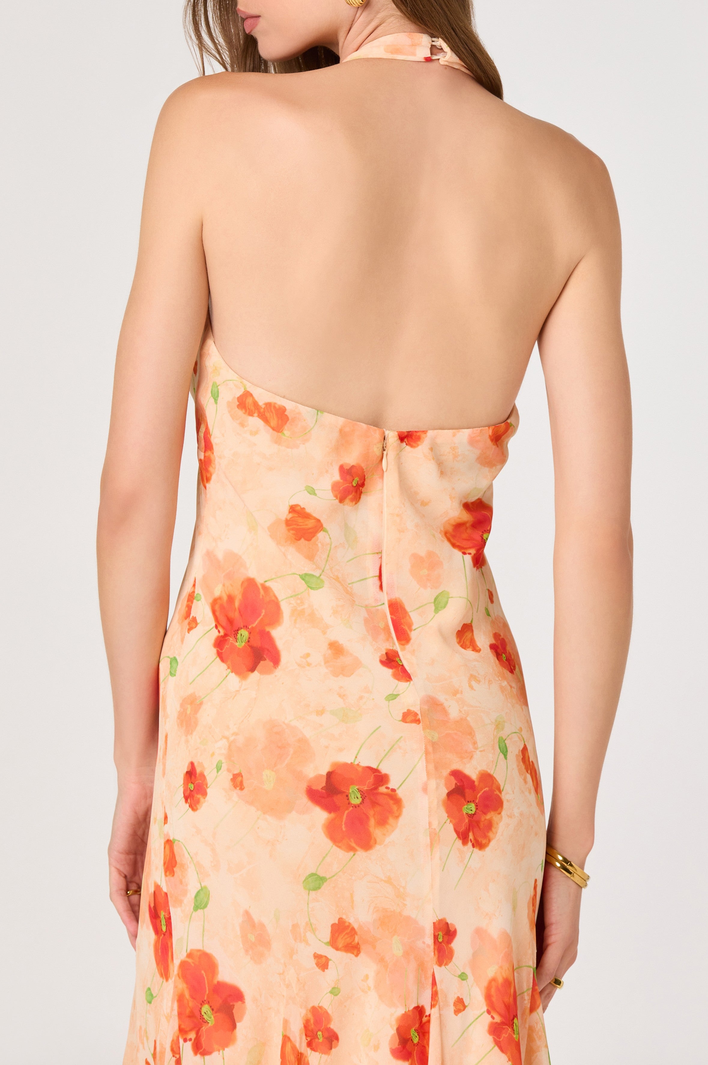 A woman stands with her back to the camera in the Yasmeen Maxi Dress by ASTR, featuring pleated straps, a backless halter design, and a peach and red floral chiffon fabric.