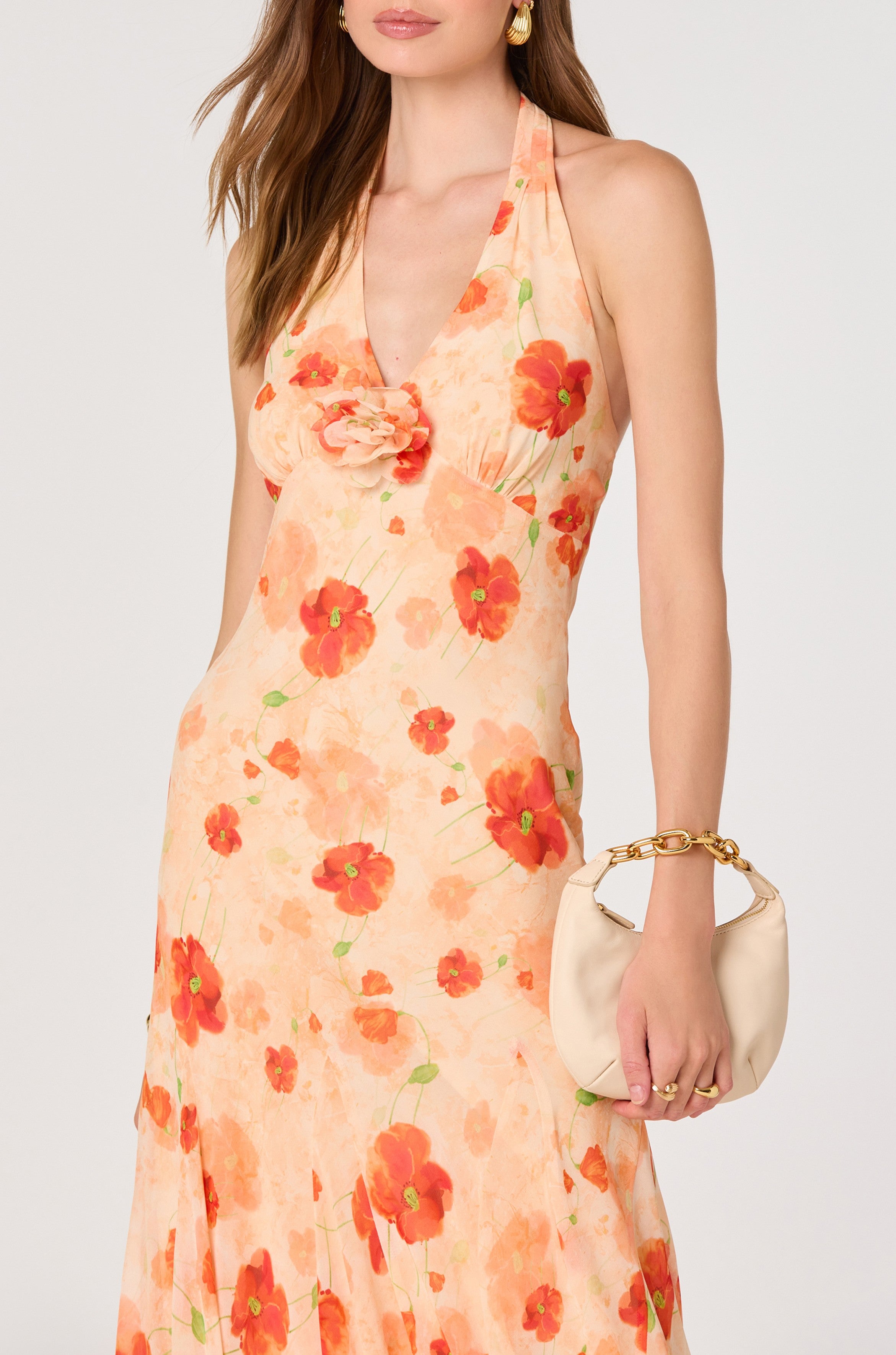 A woman in the ASTR Yasmeen Maxi Dress—an orange floral, sleeveless chiffon maxi with a halter V-neck—holds a cream handbag with a gold handle. She wears her hair loose and accessorizes with gold earrings.