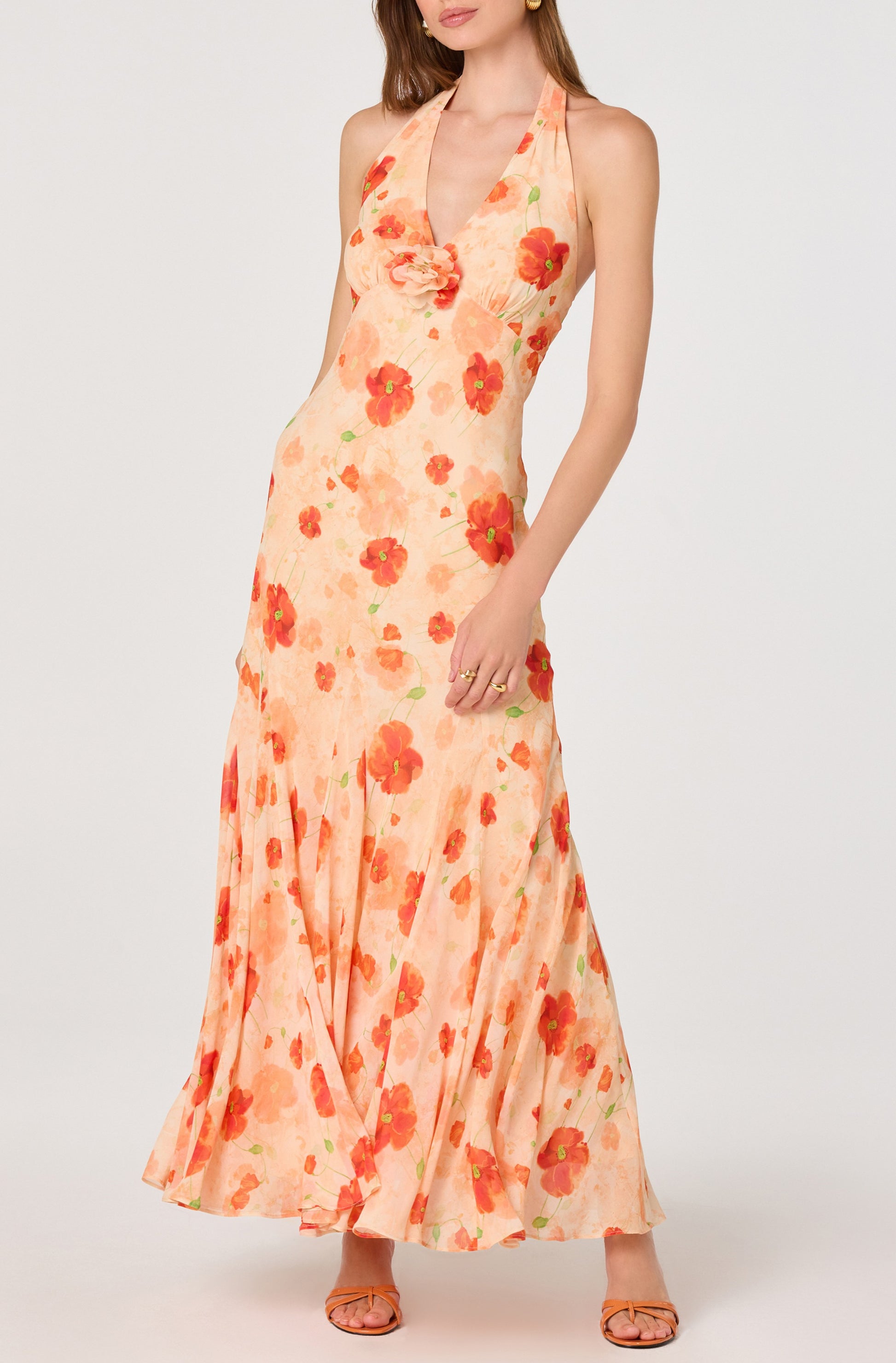 A woman in the ASTR Yasmeen Maxi Dress—peach chiffon with red and orange floral print, pleated straps, and a halter V neckline—pairs it with matching open-toe sandals against a plain white background.