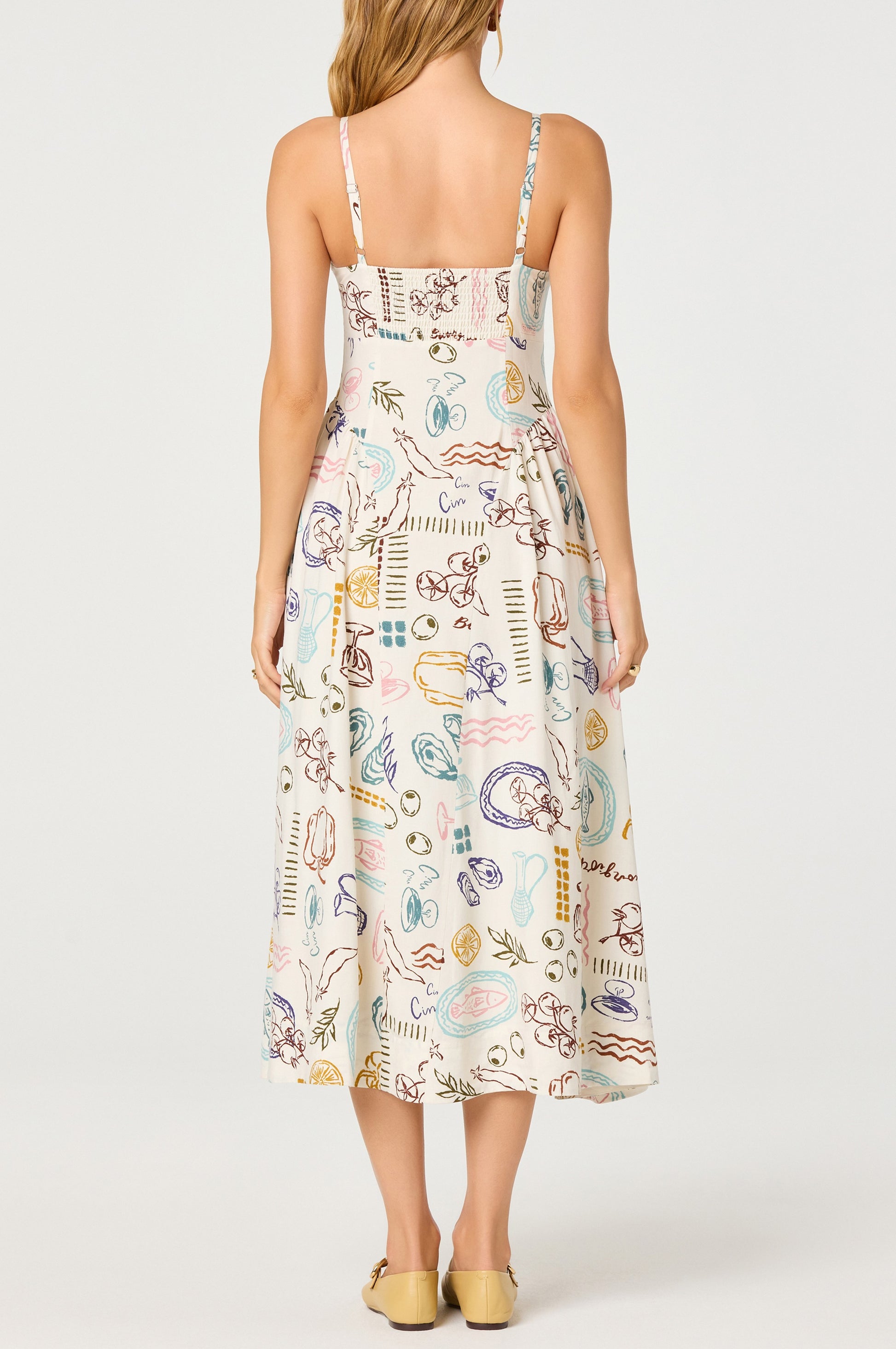 A woman faces away, wearing the ASTR Ansley Midi Dress—a colorful abstract-print linen dress with adjustable straps. The sleeveless, ankle-length style is paired with tan closed-toe shoes, her long wavy hair against a plain white background.