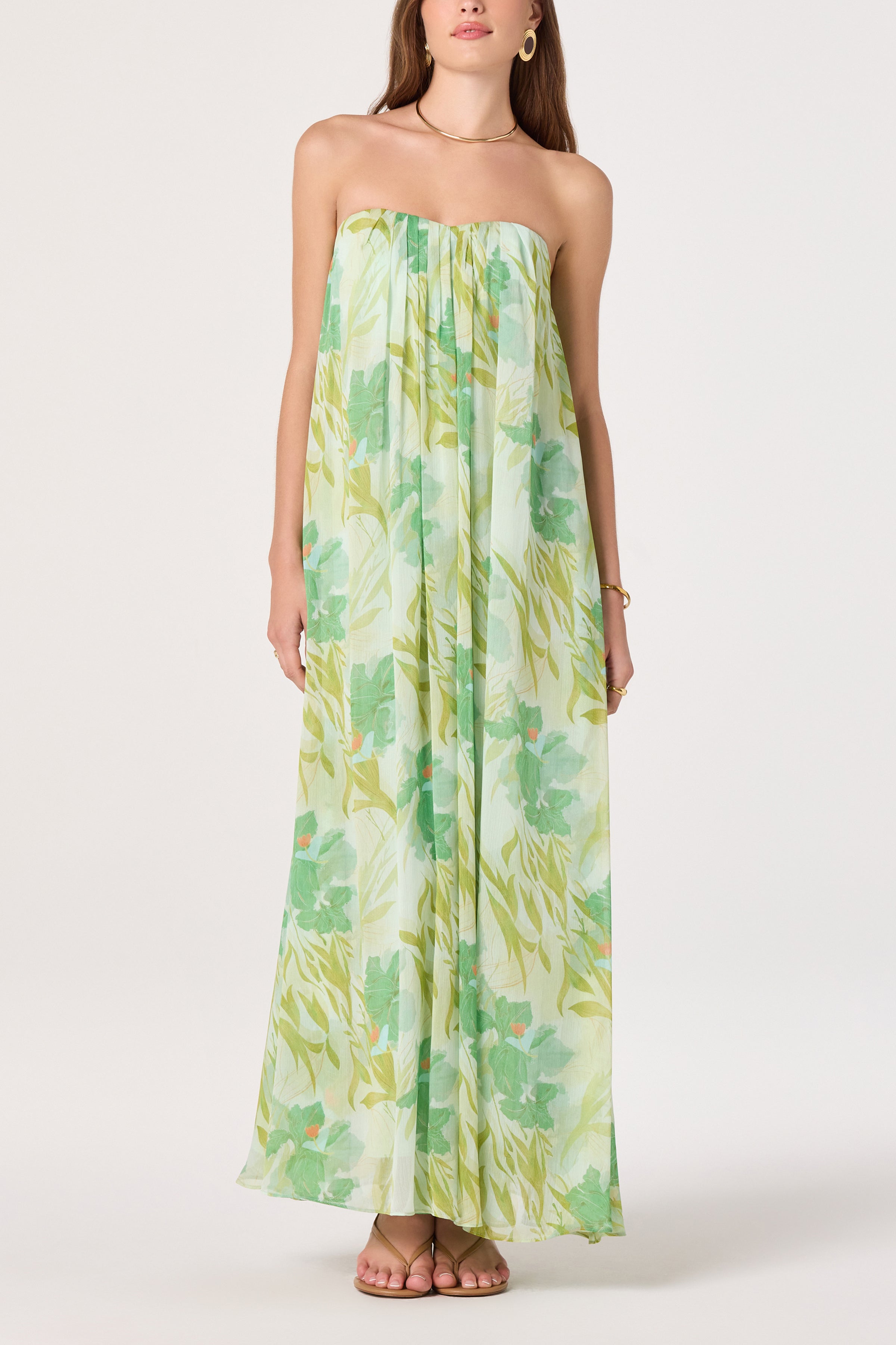 A woman models the Aileen Dress by ASTR in Green Multi, a strapless chiffon maxi with a floral print. She stands before a white background, styled with hoop earrings, a bracelet, and sandals.