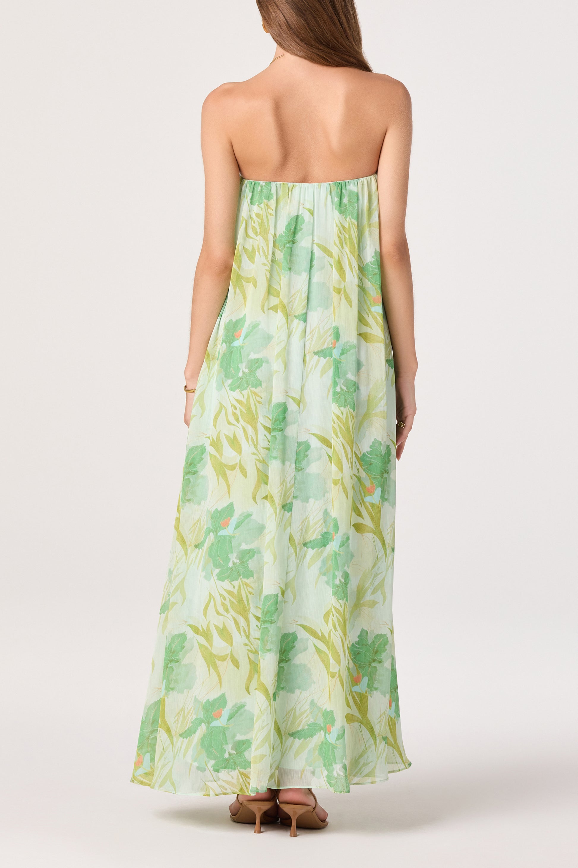 A woman with long brown hair and nude heels faces away, wearing the ASTR Aileen Dress—a flowy, strapless chiffon maxi with a green and yellow floral print—set against a white background.