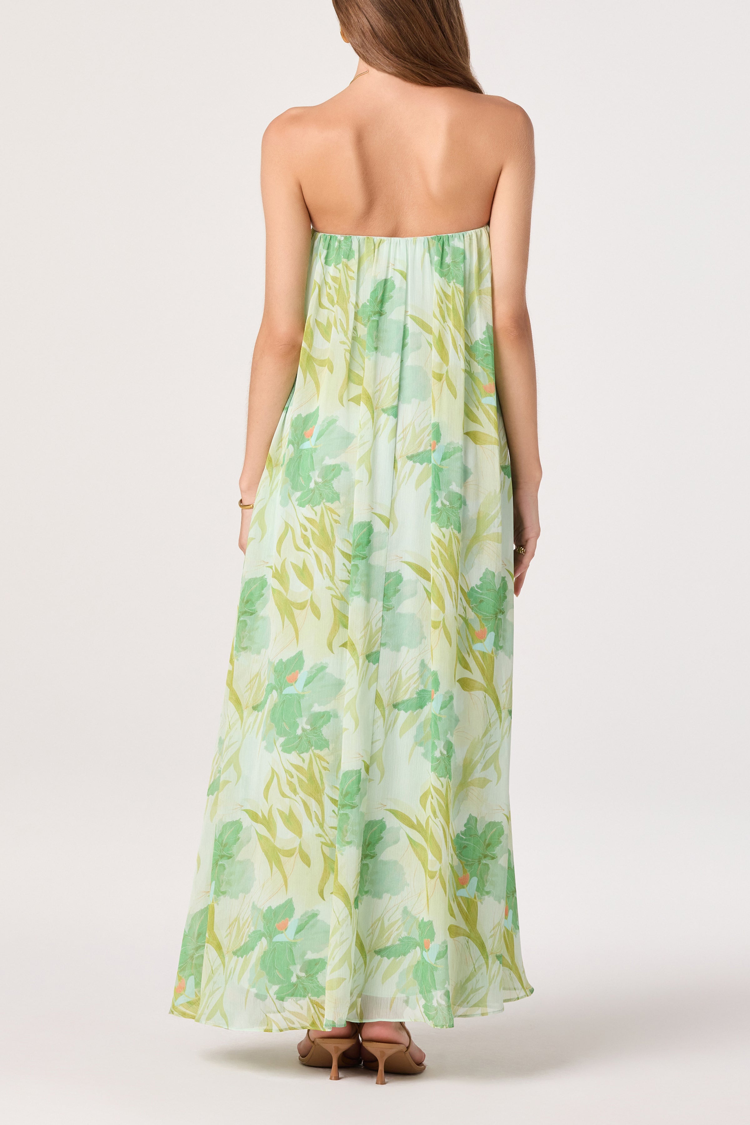 A woman with long brown hair and nude heels faces away, wearing the ASTR Aileen Dress—a flowy, strapless chiffon maxi with a green and yellow floral print—set against a white background.