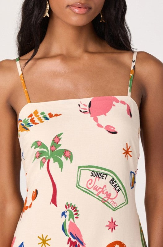 A woman wears the ASTR Valencia Dress, a cream-colored linen mini with colorful tropical prints—crab, palm tree, parrot, and "Sunset Beach Surfing"—featuring adjustable straps and an open back. Only her upper body is visible.