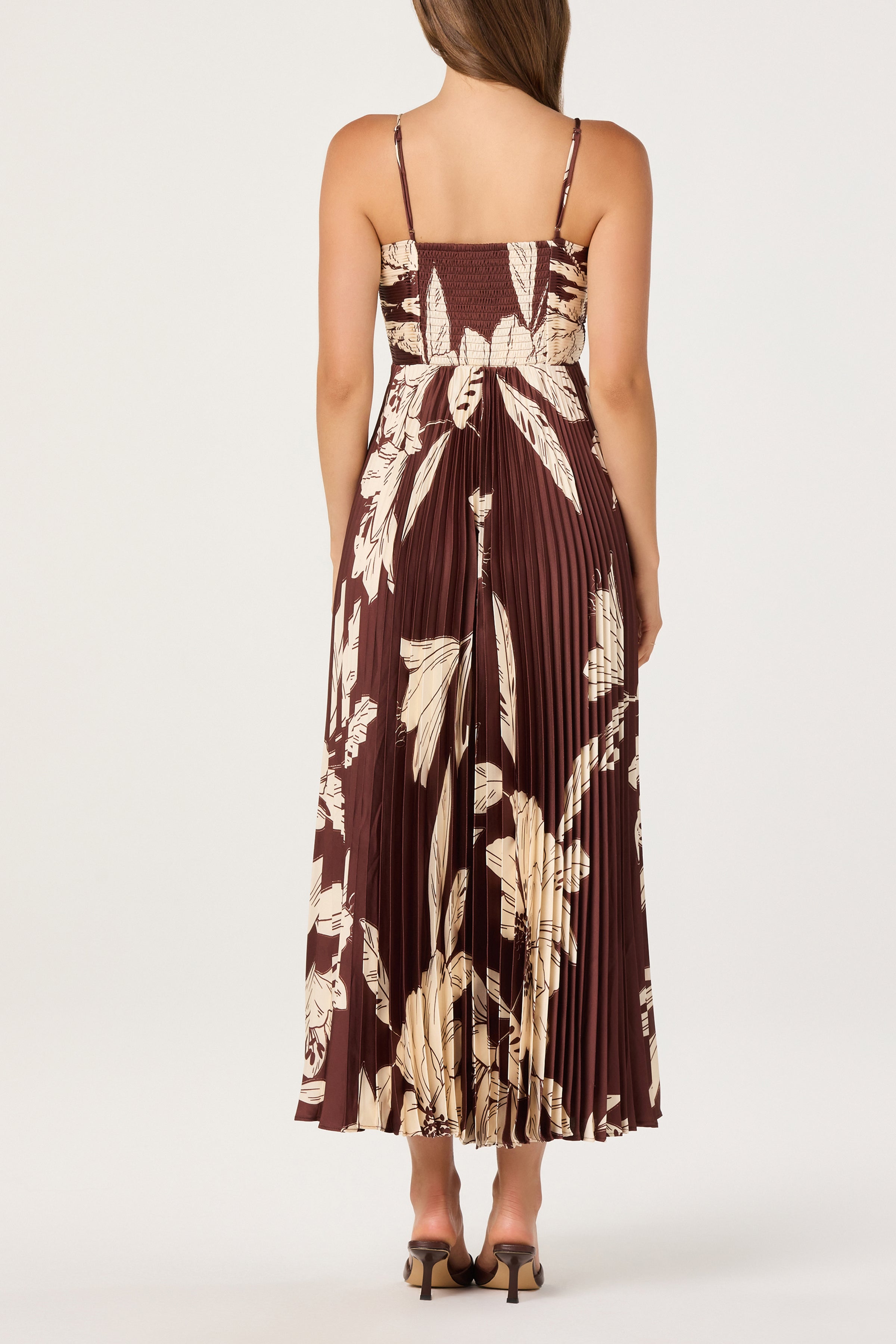 A woman faces away, wearing the ASTR Clea Dress—a brown plissé dress with beige florals, adjustable spaghetti straps, and a smocked back. She pairs it with open heels against a plain light background.