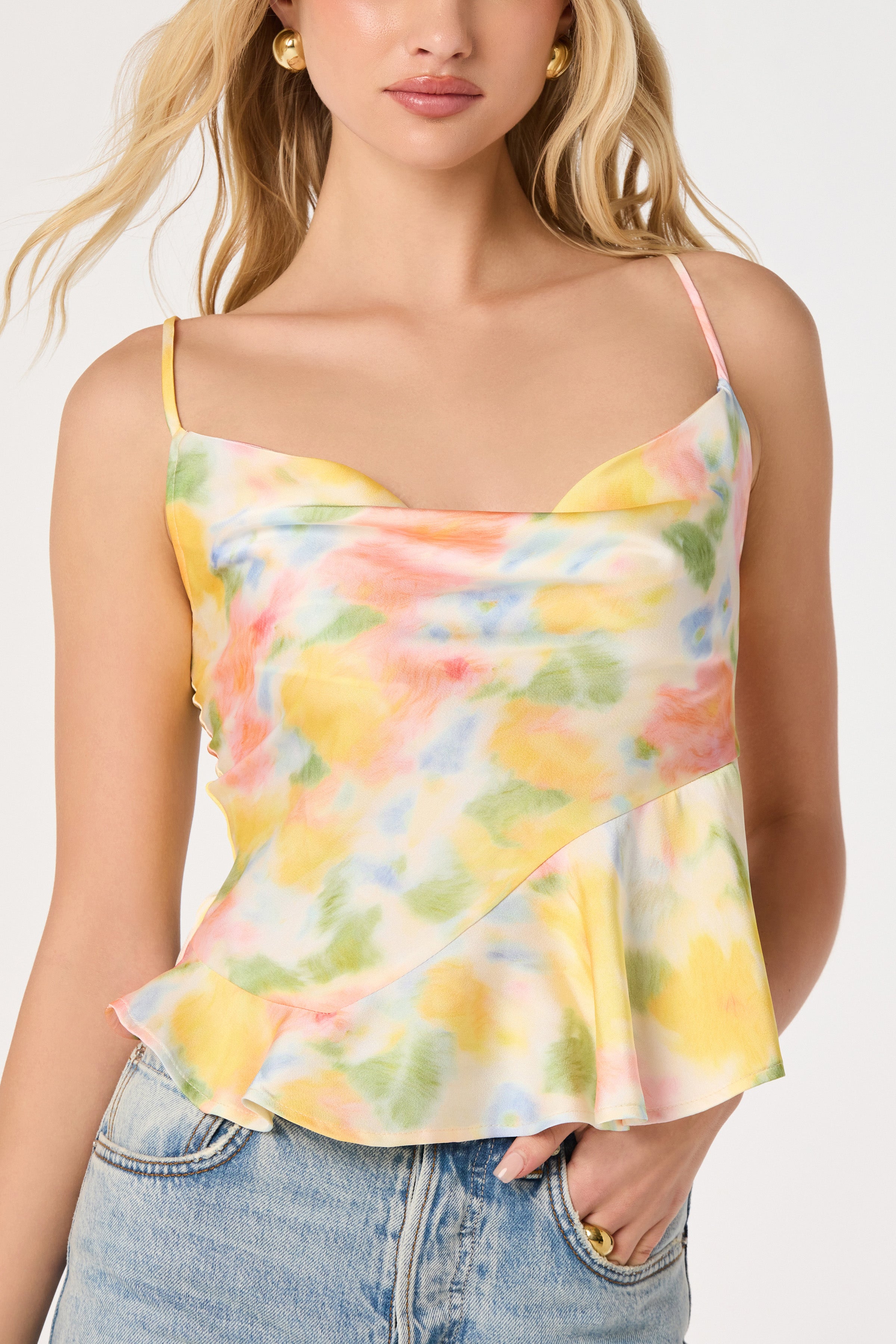 A woman wears the Everli Top by ASTR in Pink Yellow Floral, a sleeveless camisole with a cowl neckline and slight peplum hem, paired with light blue denim jeans.