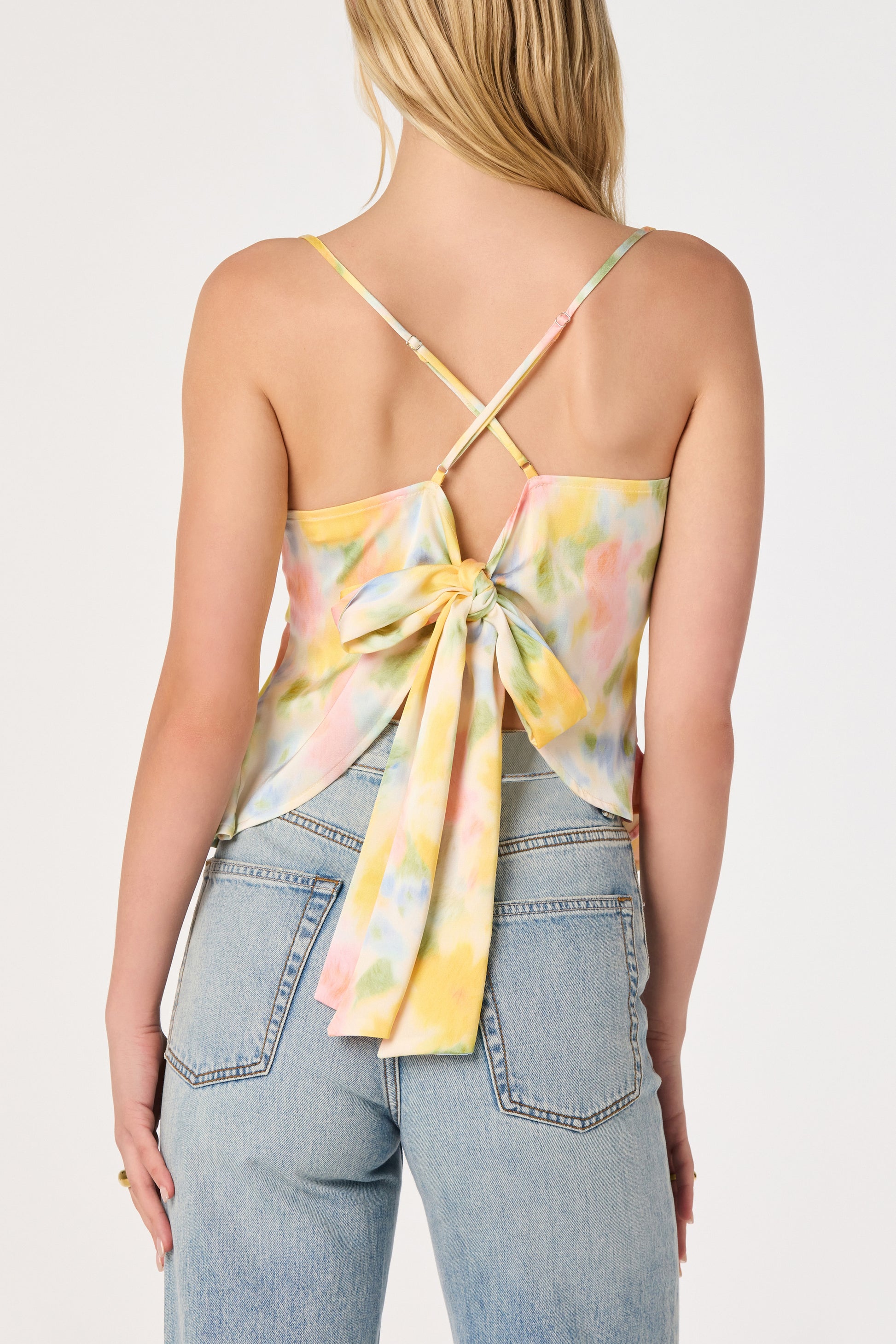 A woman, seen from behind, wears the ASTR Everli Top—a pastel tie-dye floral cami with crisscross straps and a large back bow—paired with light blue high-waisted jeans.