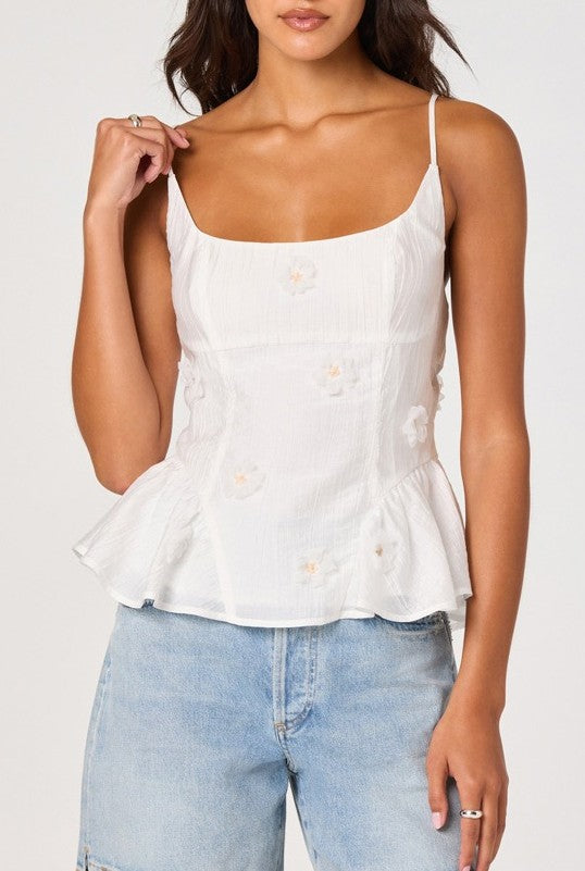 A woman wears the ASTR Kassidy Top, a sleeveless white peplum with adjustable spaghetti straps and floral embroidery, paired with light blue jeans. The image is cropped at the shoulders and thighs.
