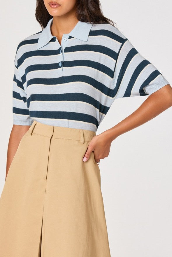 A woman is wearing the ASTR Scottie Sweater in BLUE STRIPE—a short-sleeve, lightweight knit polo with a ribbed collar—tucked into high-waisted beige pants, with one hand on her hip.