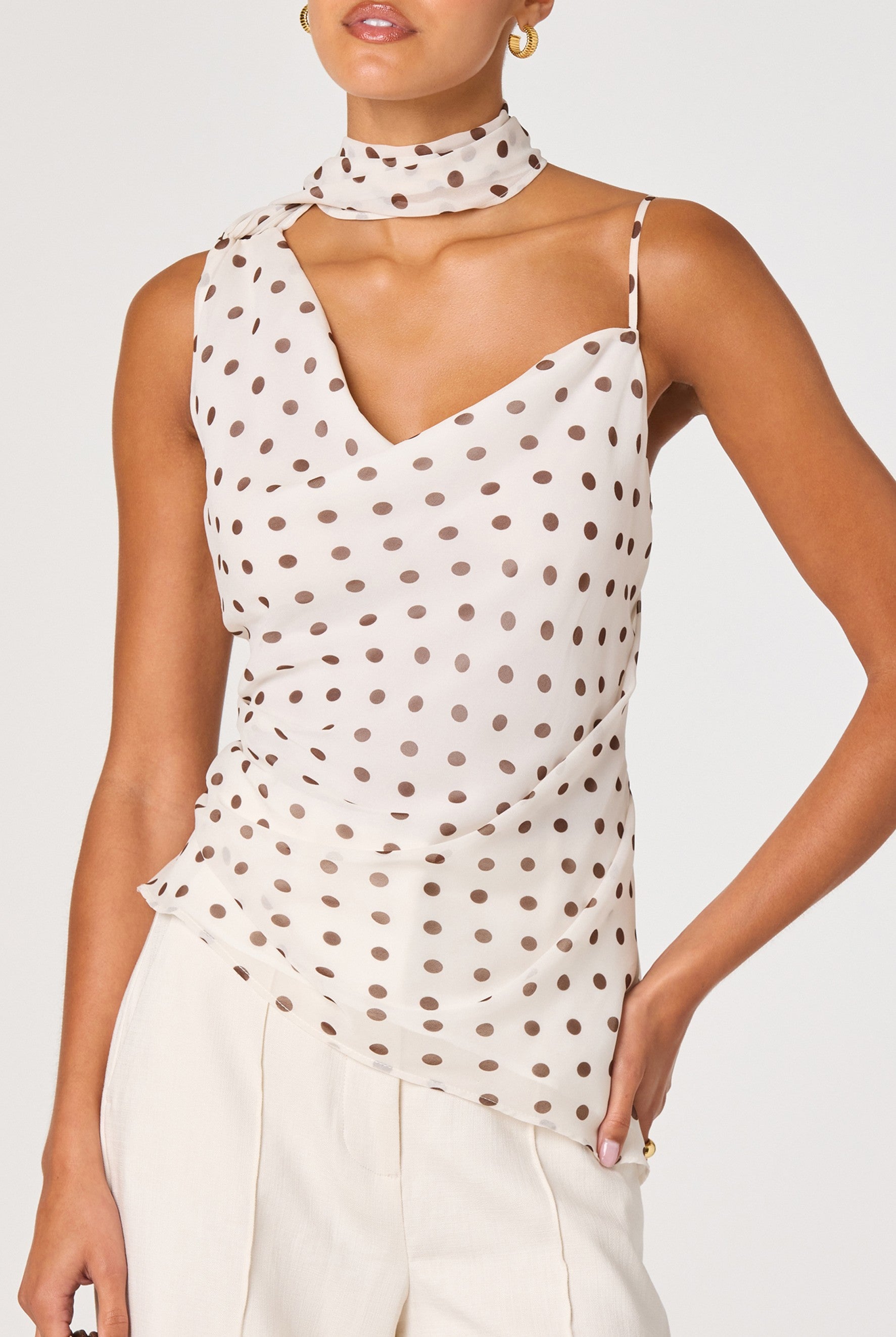 A woman wears the ASTR Adrie Top—an ivory chiffon piece with brown polka dots, an asymmetrical cowl neckline, adjustable straps, and a matching scarf—paired with light-colored trousers.