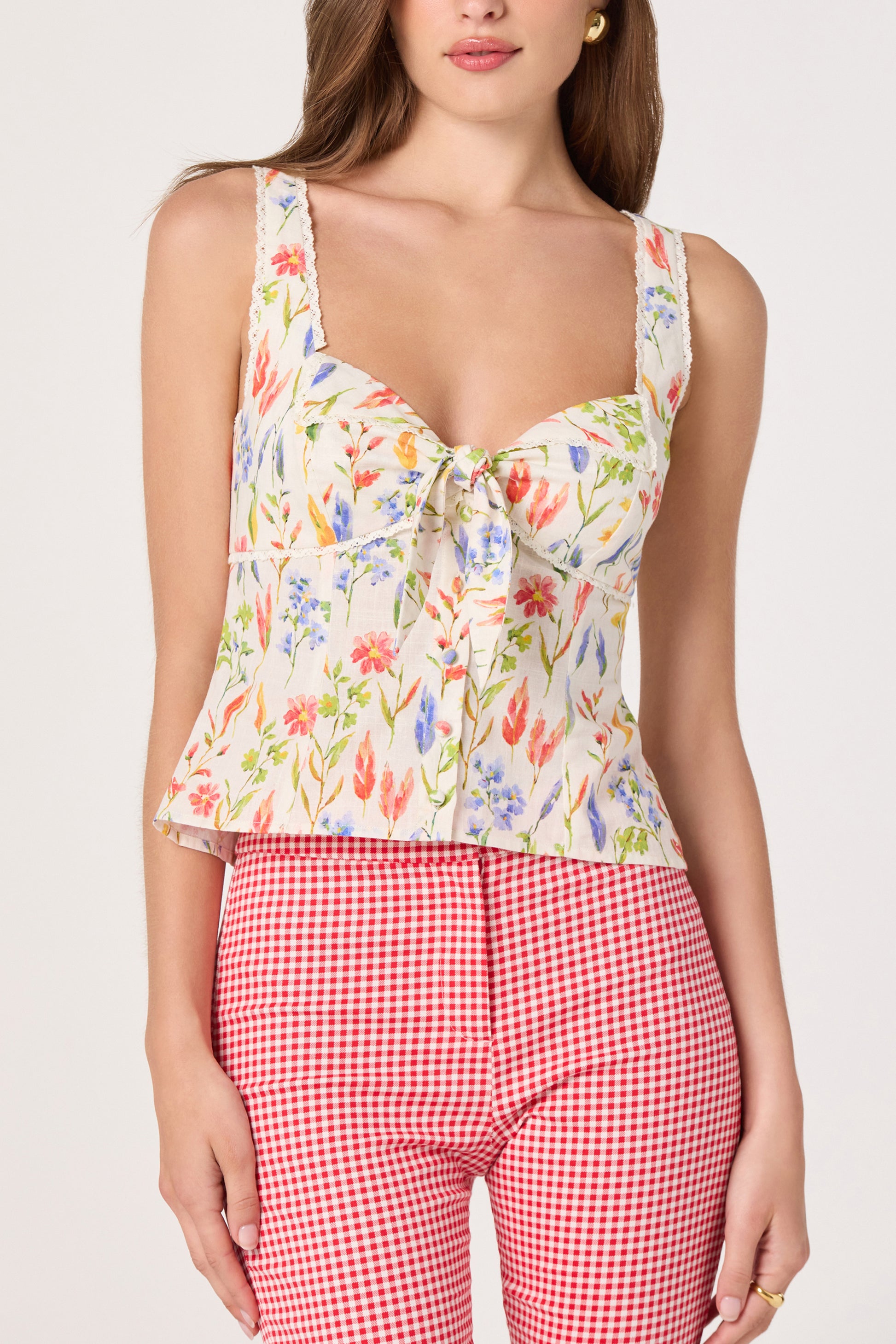 A woman wears the ASTR Theresa Top in White Multi—a slim fit, floral sleeveless top with a square neckline and front tie—paired with high-waisted red and white checkered pants against a plain white background.