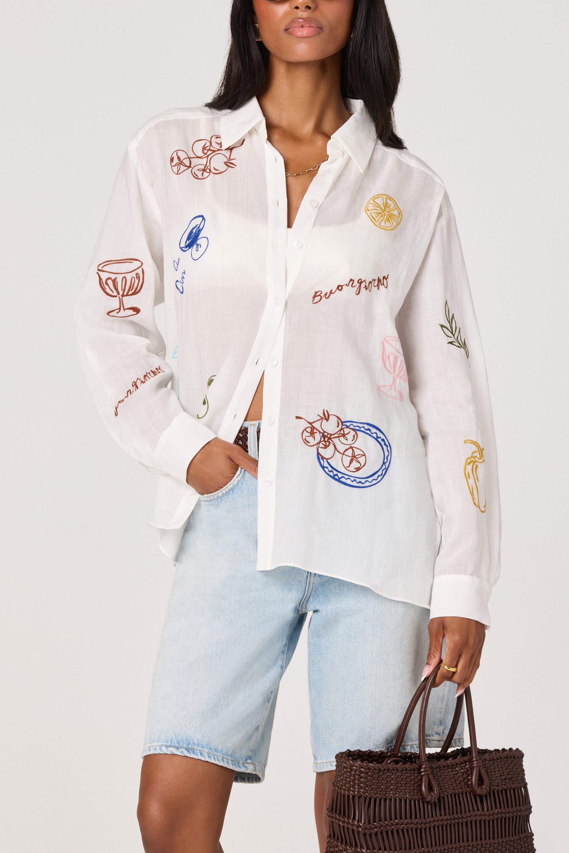 A woman wearing the ASTR Trace Shirt—a white embroidered top with colorful designs—paired with light denim Bermuda shorts and carrying a dark brown woven handbag.