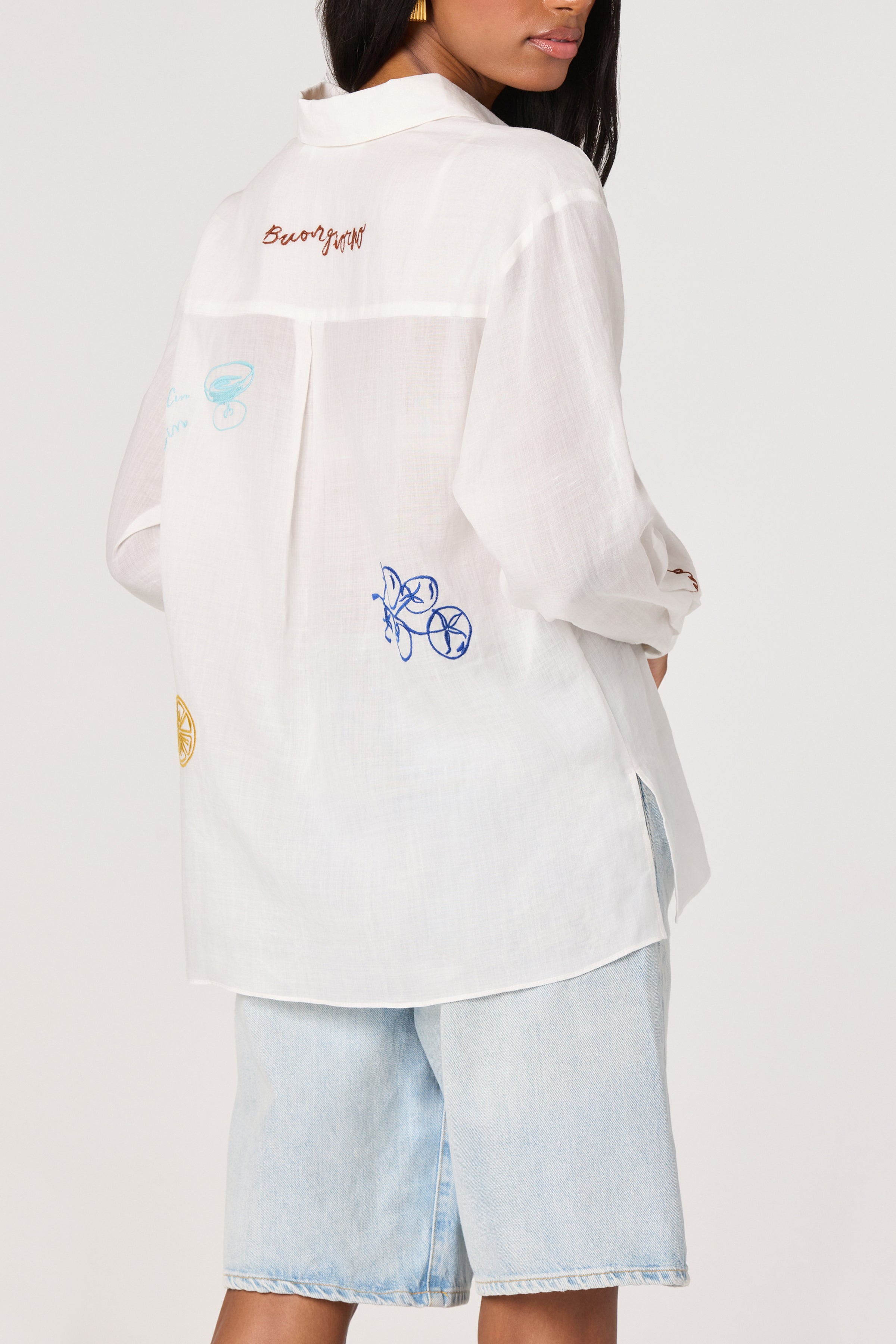 A woman wearing the ASTR Trace Shirt, a loose semi-sheer top with colorful embroidery on the back, paired with light blue denim shorts, stands turned away against a plain background.