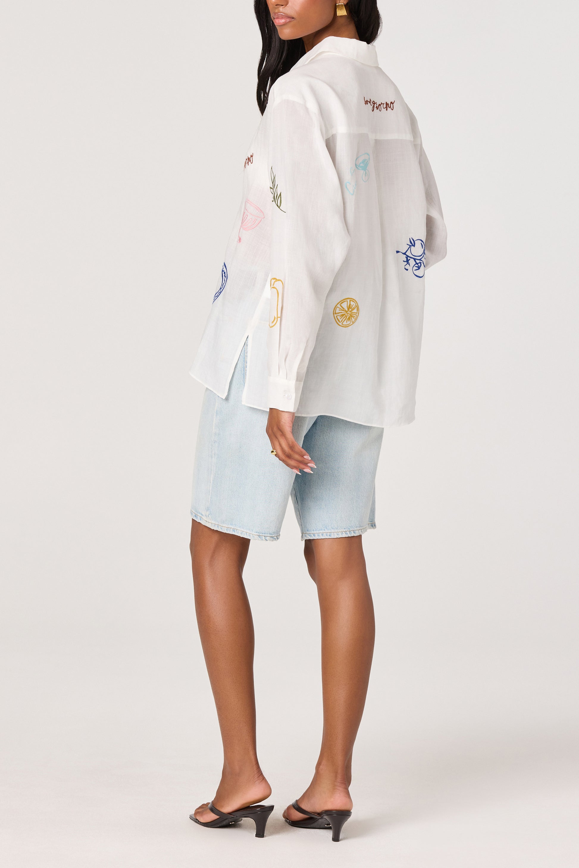 A woman wearing the ASTR Trace Shirt with colorful embroidery, light denim knee-length shorts, and black heeled sandals stands with her back turned and head slightly to the side.