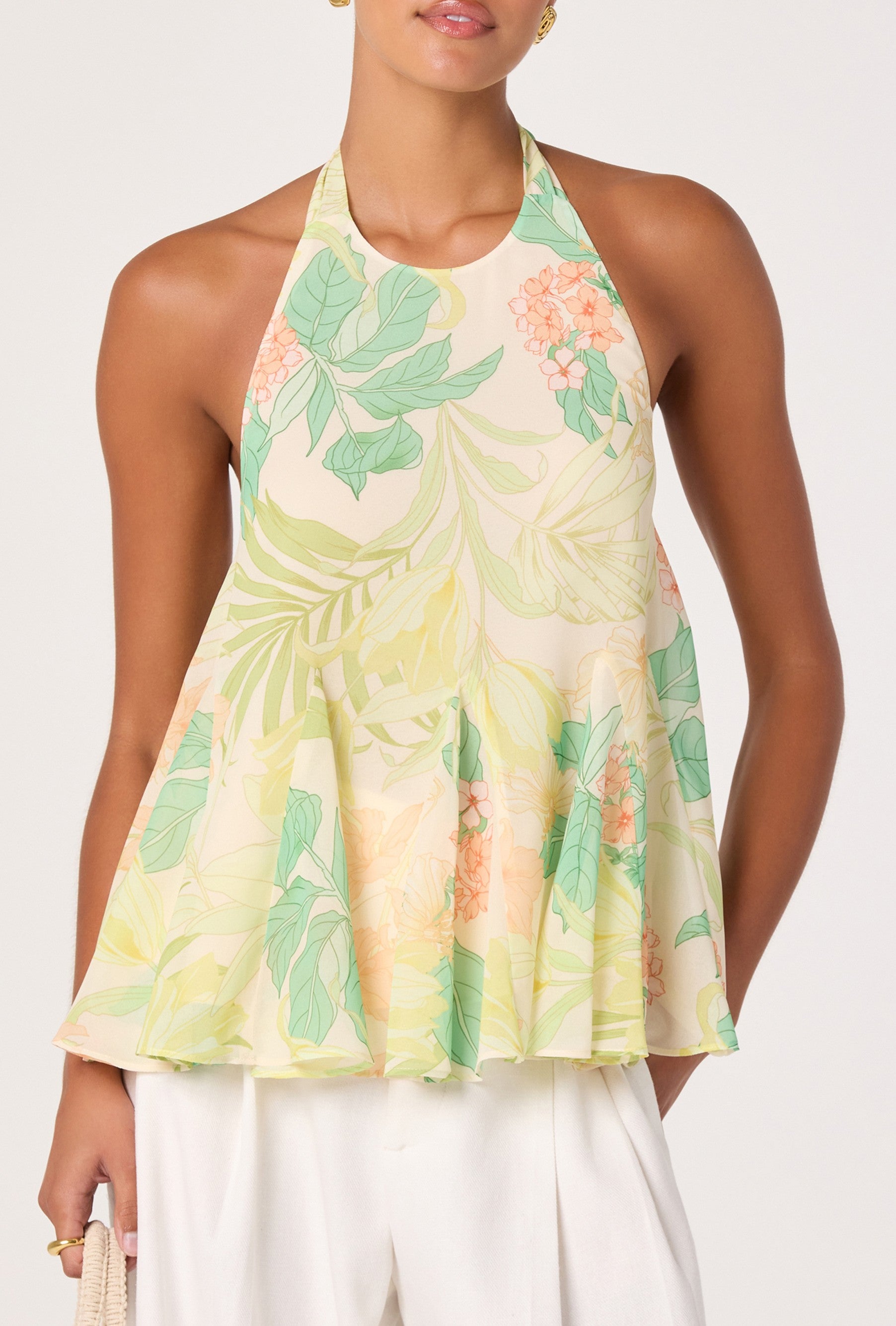 A woman wears the ASTR Belen Top, a sleeveless chiffon halter with a green multi floral print, paired with white pants and a woven bag. The top features an open back. The background is light and plain.