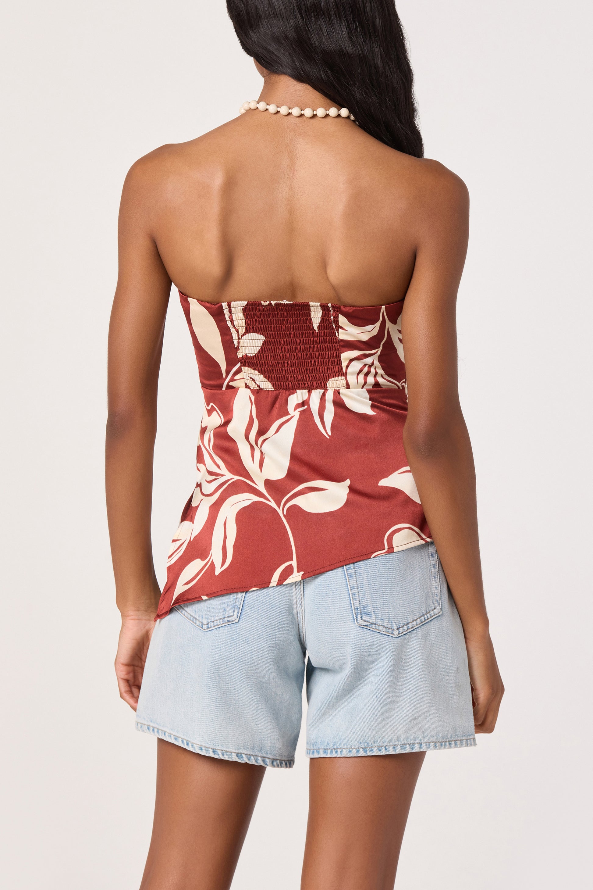 A woman stands facing away, wearing the ASTR Annabeth Top—a satin strapless piece with a red and white floral pattern—paired with light blue denim shorts and a beaded necklace. The top’s asymmetric hem adds a modern twist to her look, while her long hair drapes over one shoulder.