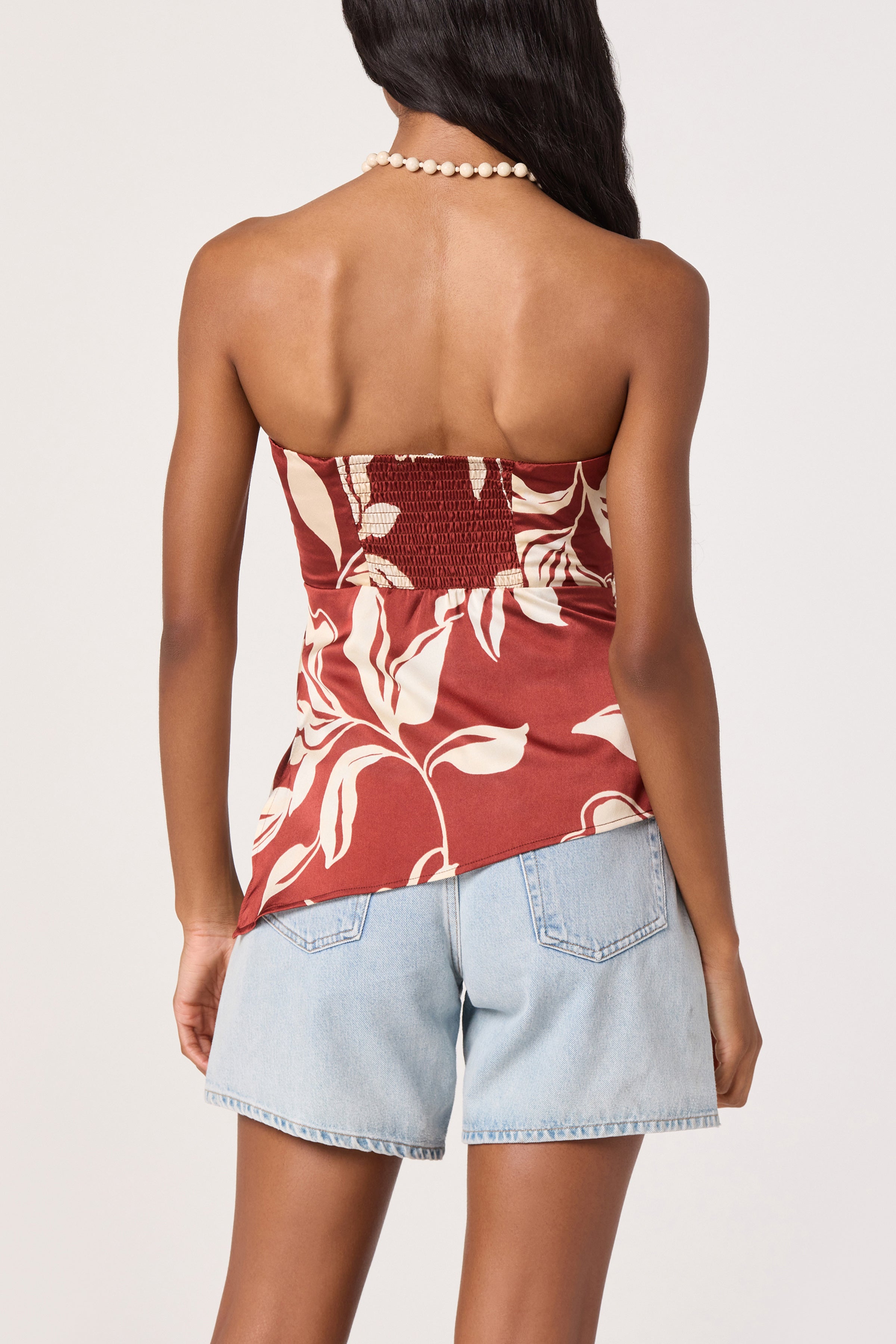 A woman stands facing away, wearing the ASTR Annabeth Top—a satin strapless piece with a red and white floral pattern—paired with light blue denim shorts and a beaded necklace. The top’s asymmetric hem adds a modern twist to her look, while her long hair drapes over one shoulder.