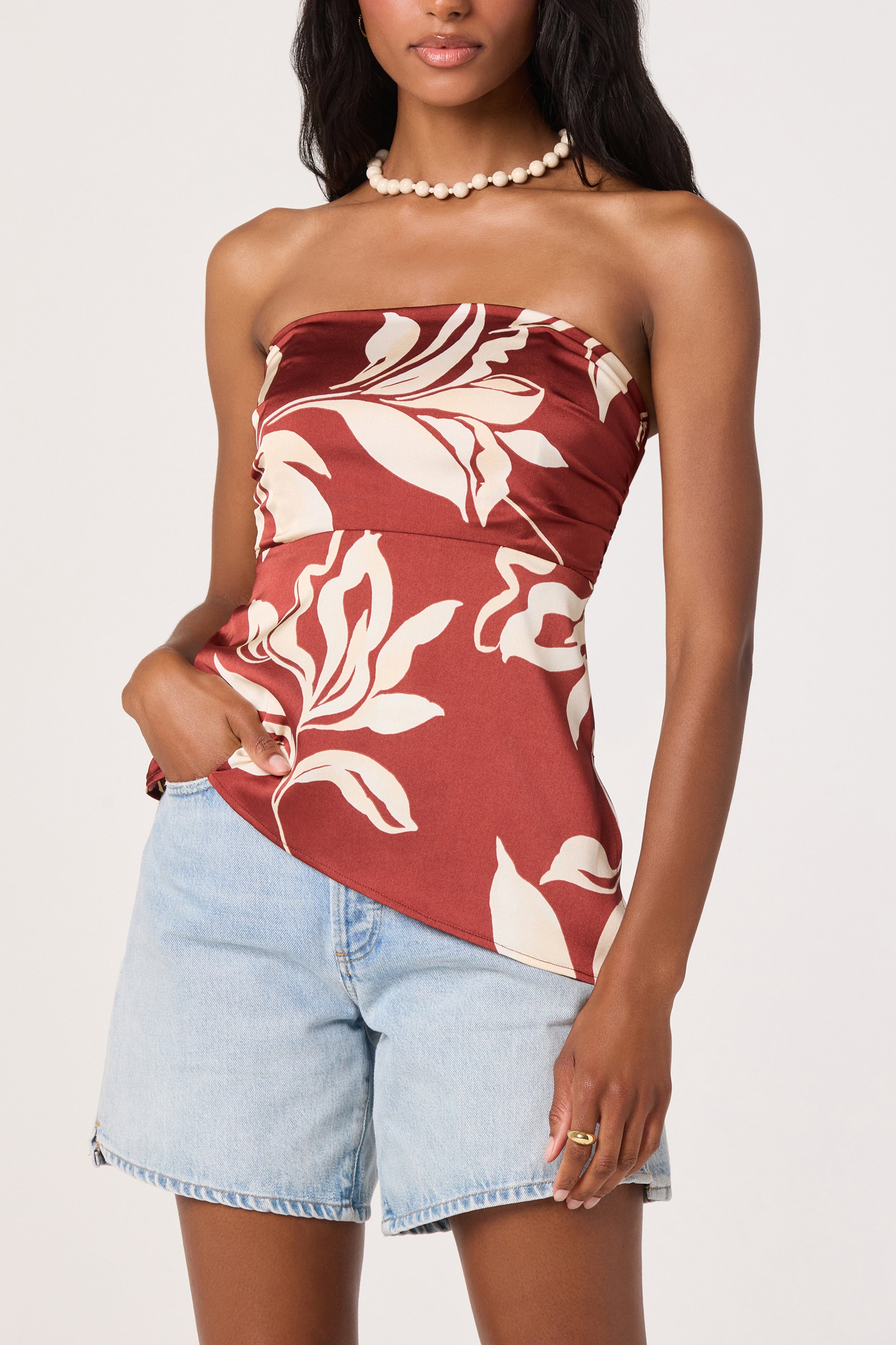 A woman wearing the ASTR Annabeth Top in Brown Floral—a ruched bust, satin strapless top—paired with light blue denim shorts, a pearl necklace, and a ring, standing with one hand in her pocket.