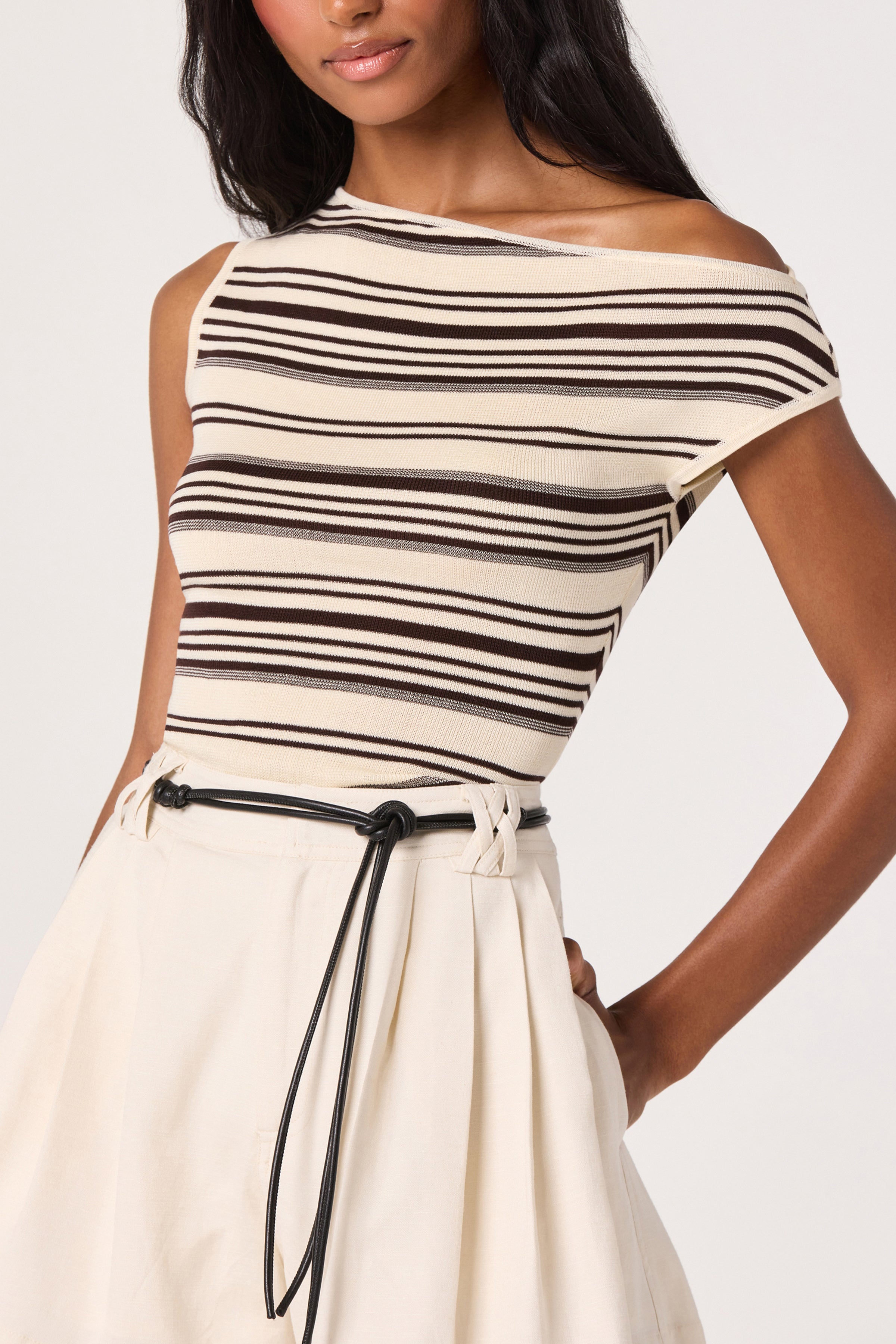 A woman is wearing the Sofia Top by ASTR, an off-the-shoulder, sleeveless beige fitted knit top featuring black diagonal stripes, styled with a high-waisted cream skirt and a thin black belt. Only her upper body is visible against a plain white background.