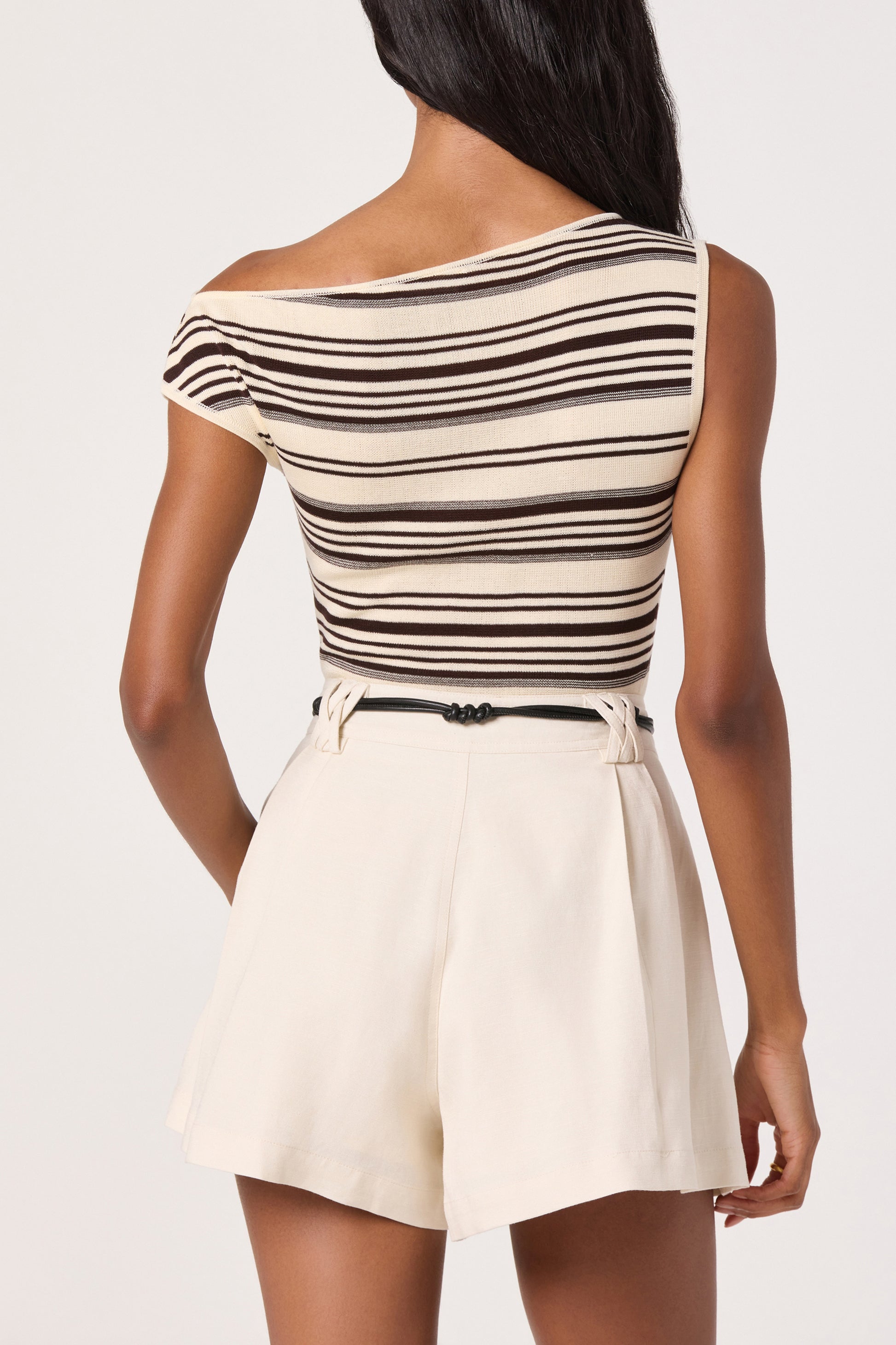 A woman stands facing away, wearing the ASTR Sofia Top—a beige and brown striped, one-shoulder viscose top—paired with high-waisted, light cream shorts and a black belt.