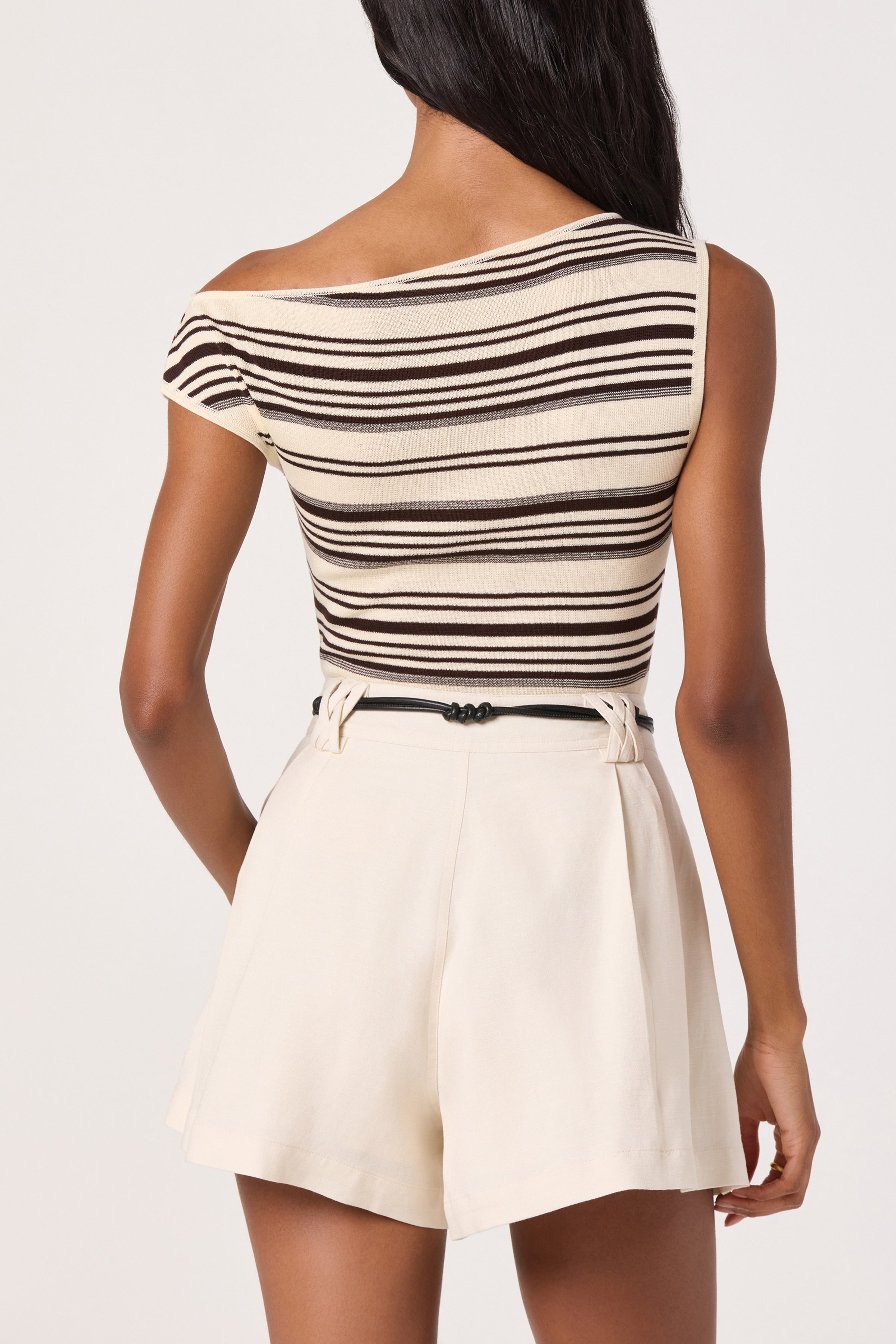 A woman stands facing away, wearing the ASTR Sofia Top—a beige and brown striped, one-shoulder viscose top—paired with high-waisted, light cream shorts and a black belt.