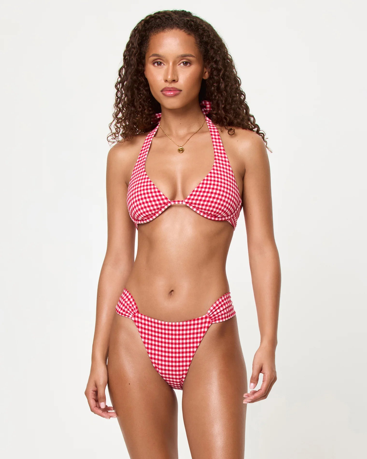 A woman with long curly hair models the LSpace Atta Girl Gingham Dolce Bottom Full in red and white, featuring ruched sides (SWBM0136), against a plain light background, looking at the camera with a neutral expression.