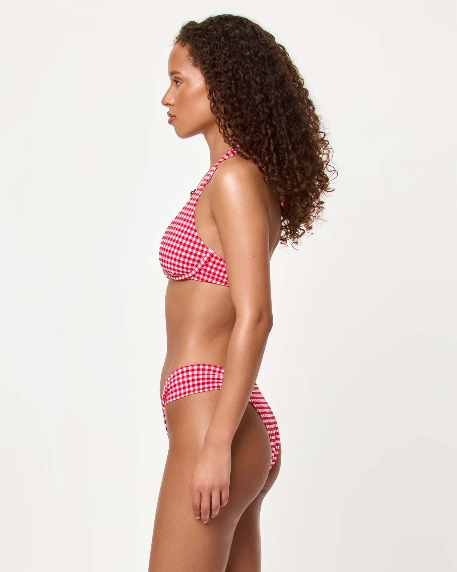 A woman with curly hair stands in profile wearing the LSpace Atta Girl Gingham Dolce Bottom Full, a mid rise red and white gingham bikini with ruched side detailing, against a plain light background.