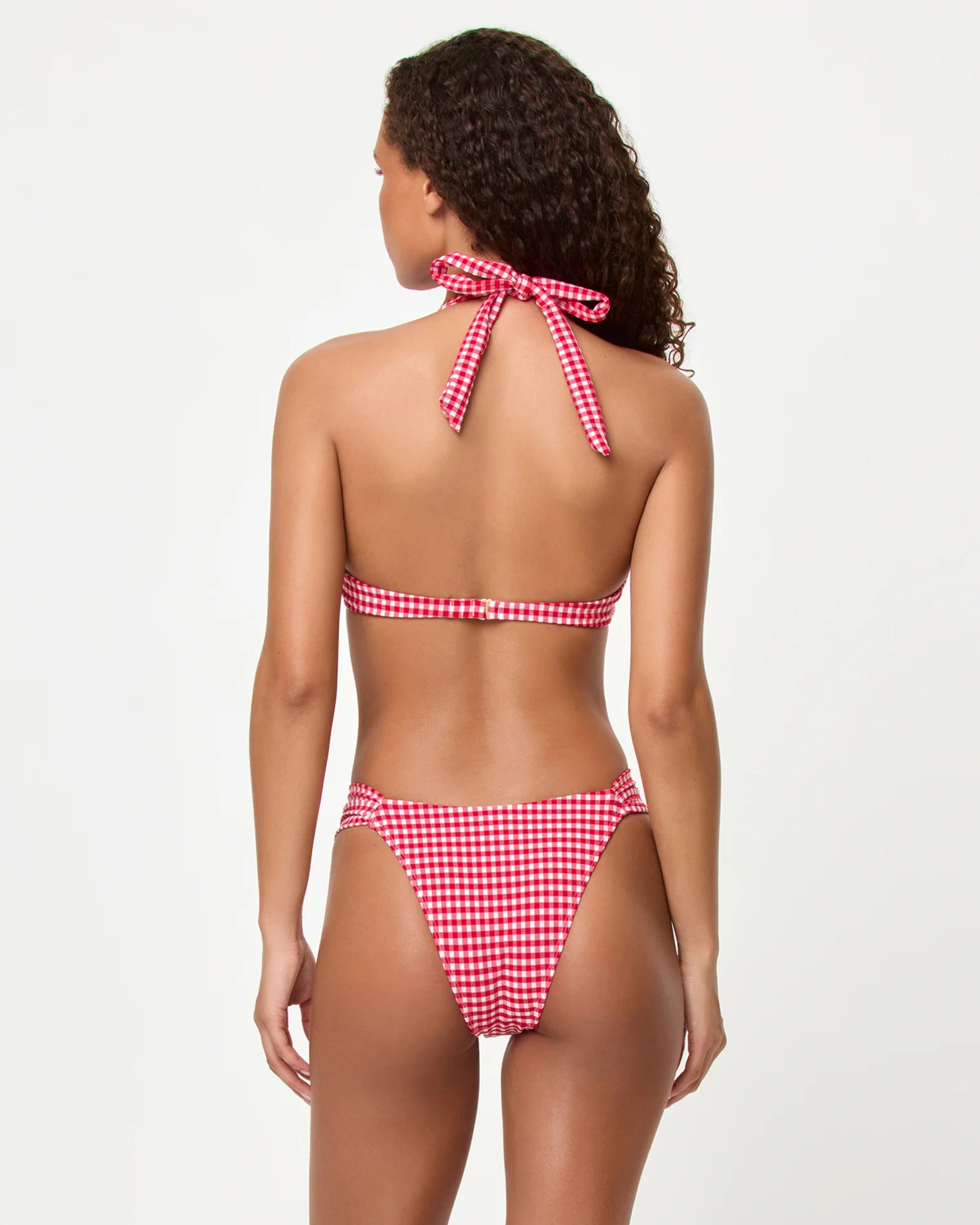 A woman with curly hair, seen from behind, wears the LSpace Atta Girl Gingham Dolce Bottom Full bikini in Red Multi against a plain light background.