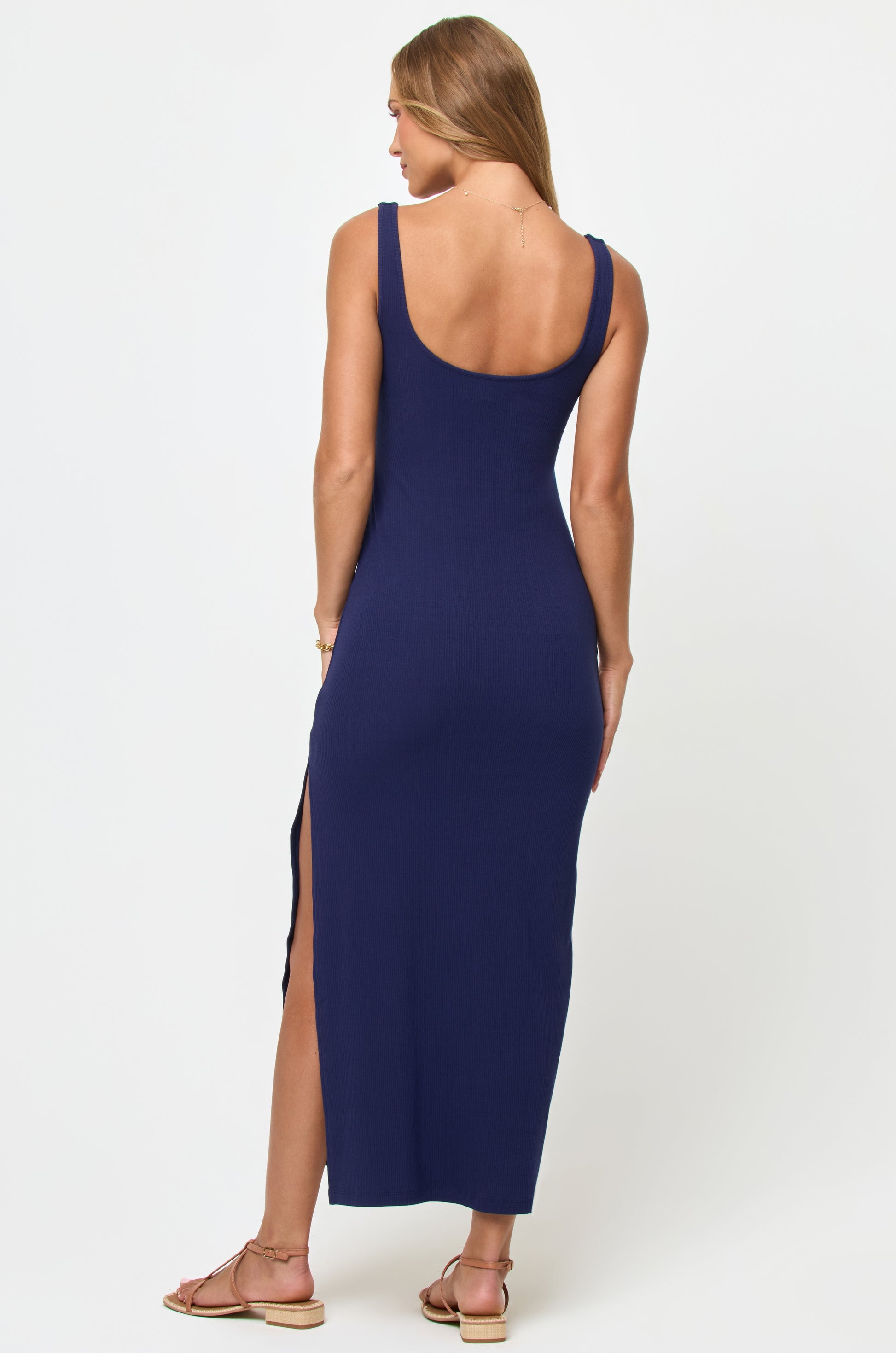A woman with long hair stands facing away against a white background, wearing the LSpace Alexis Maxi Dress in navy blue with a side leg slit and flat sandals.