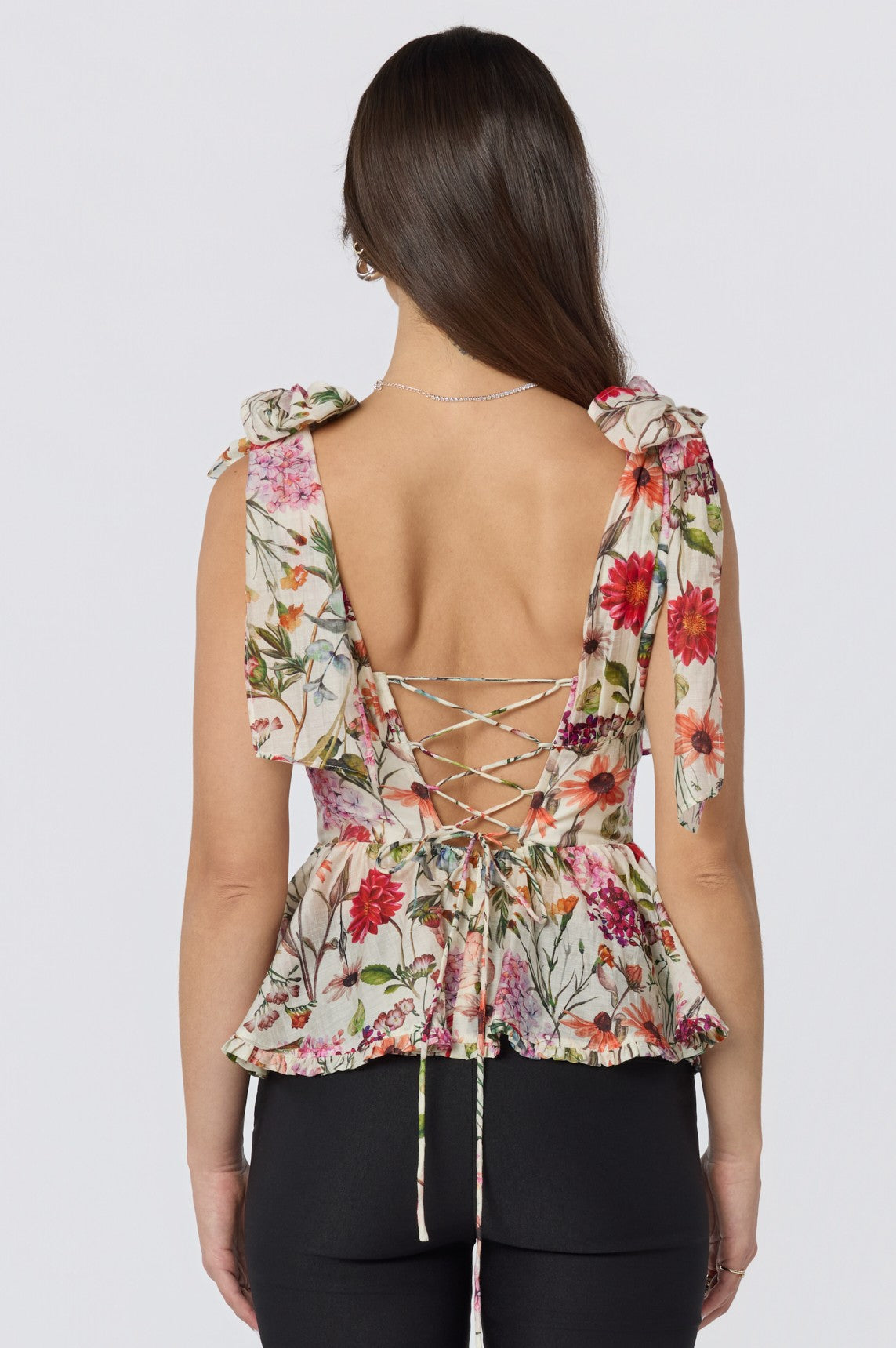 A woman seen from behind wears the ASTR Lenora Top—a floral peplum top with a sweetheart neckline, shoulder bows, lace-up back, multicolored flower pattern, and a flattering peplum hem—paired with black pants.