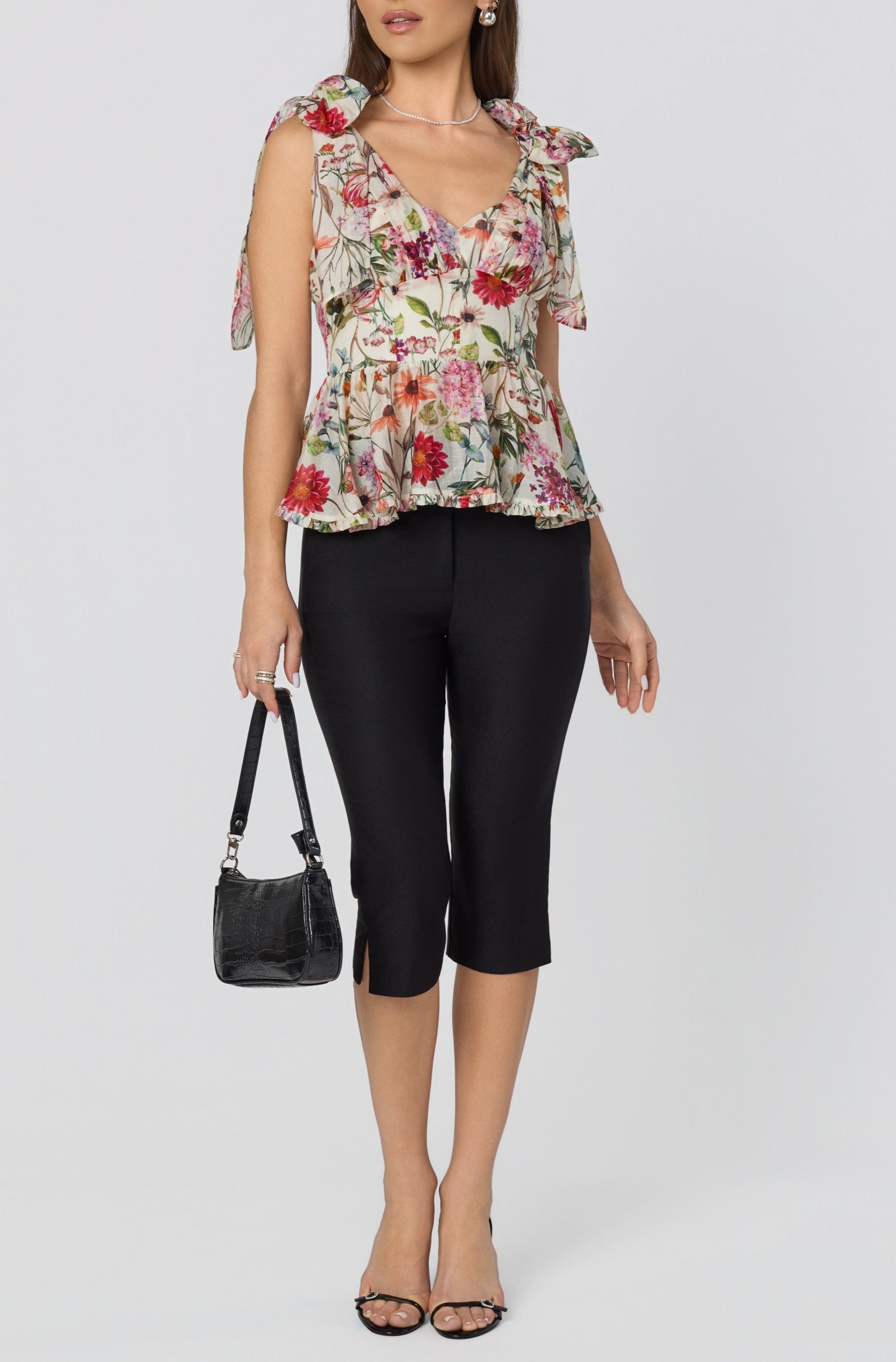 A woman wears the ASTR Lenora Top, a floral linen corset peplum with a sweetheart neckline and bow tie straps, paired with black knee-length slit pants, black heeled sandals, and holds a small black handbag against a plain background.