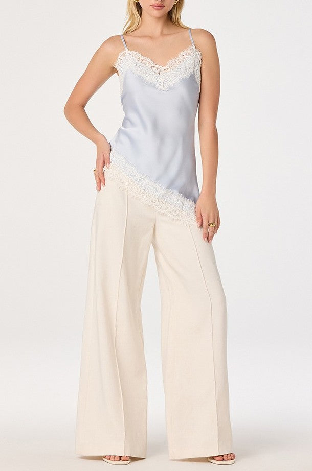 A woman wears the Analise Top by ASTR, a light blue satin tank featuring eyelash lace trim, paired with wide-leg cream pants. She poses confidently with one hand on her hip against a plain white background.
