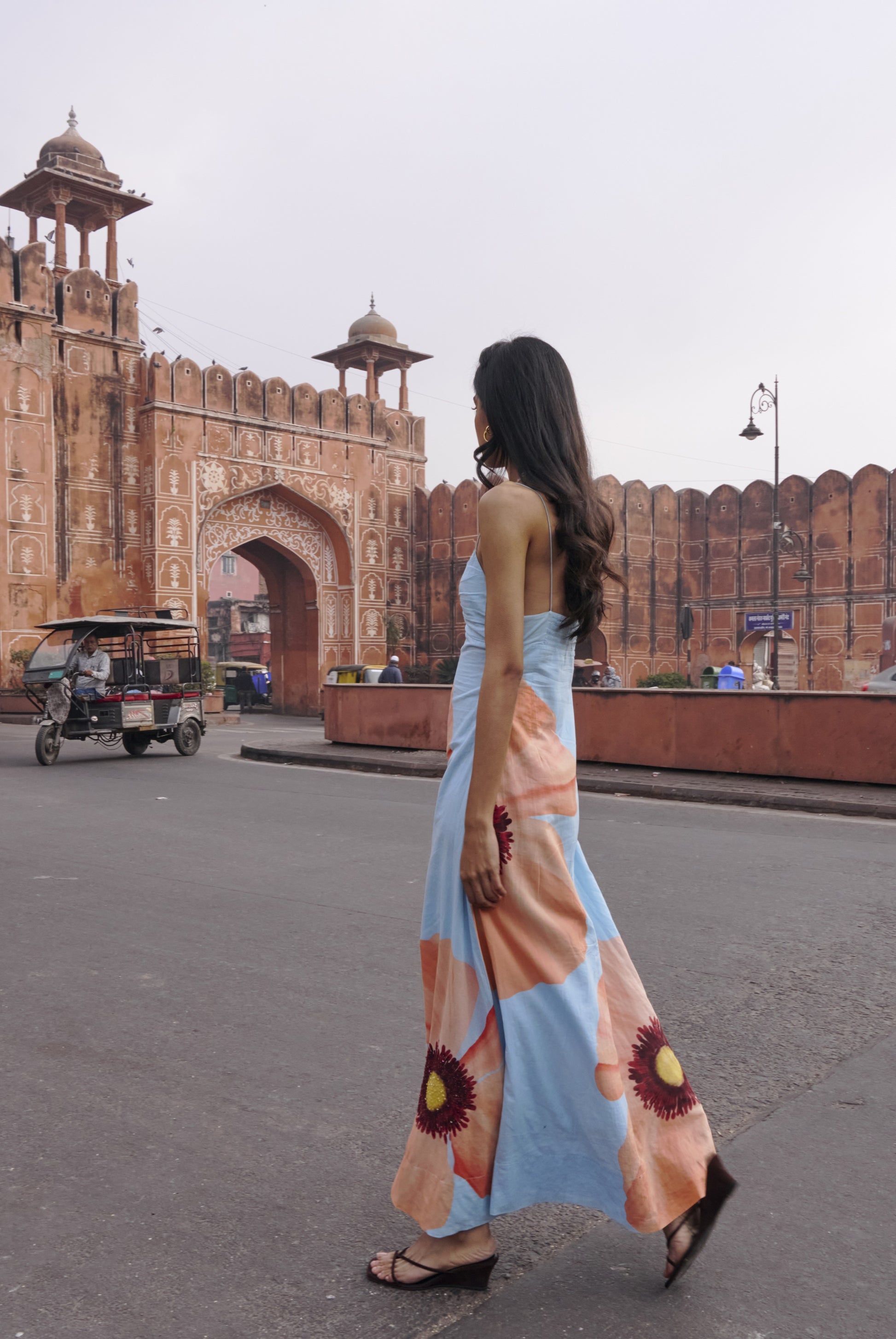 A woman in the Summer Away Alfama Maxi Dress—blue with large floral prints and adjustable straps—walks near a historic red sandstone gate, with an auto rickshaw and people in the background.