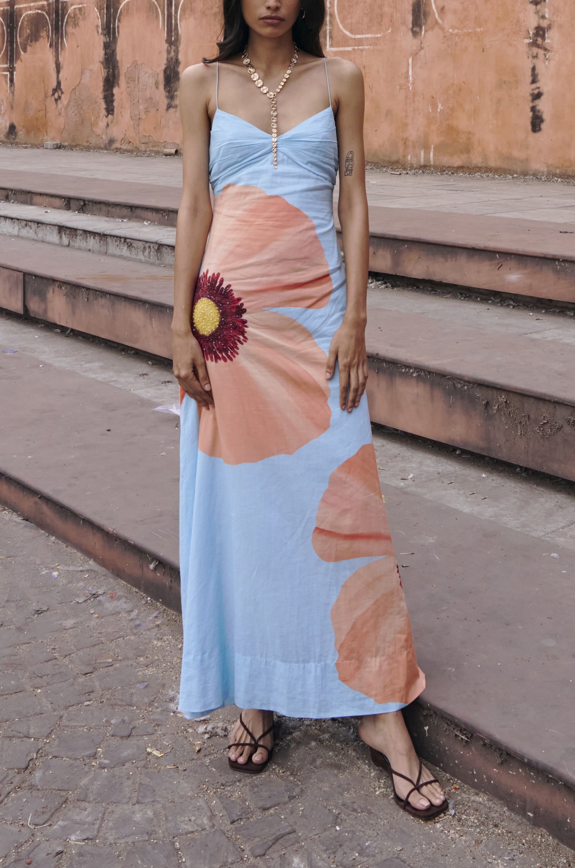 A woman in the Summer Away Alfama Maxi Dress, featuring large peach and red florals and adjustable straps, stands on stone steps before a textured peach wall.