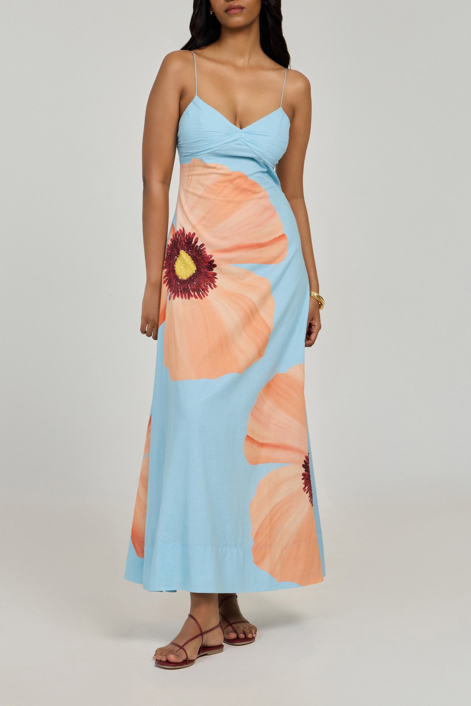 A woman wearing the Summer Away Alfama Maxi Dress—light blue, sleeveless, with adjustable straps and bold orange poppy prints—stands against a plain light background in brown sandals.