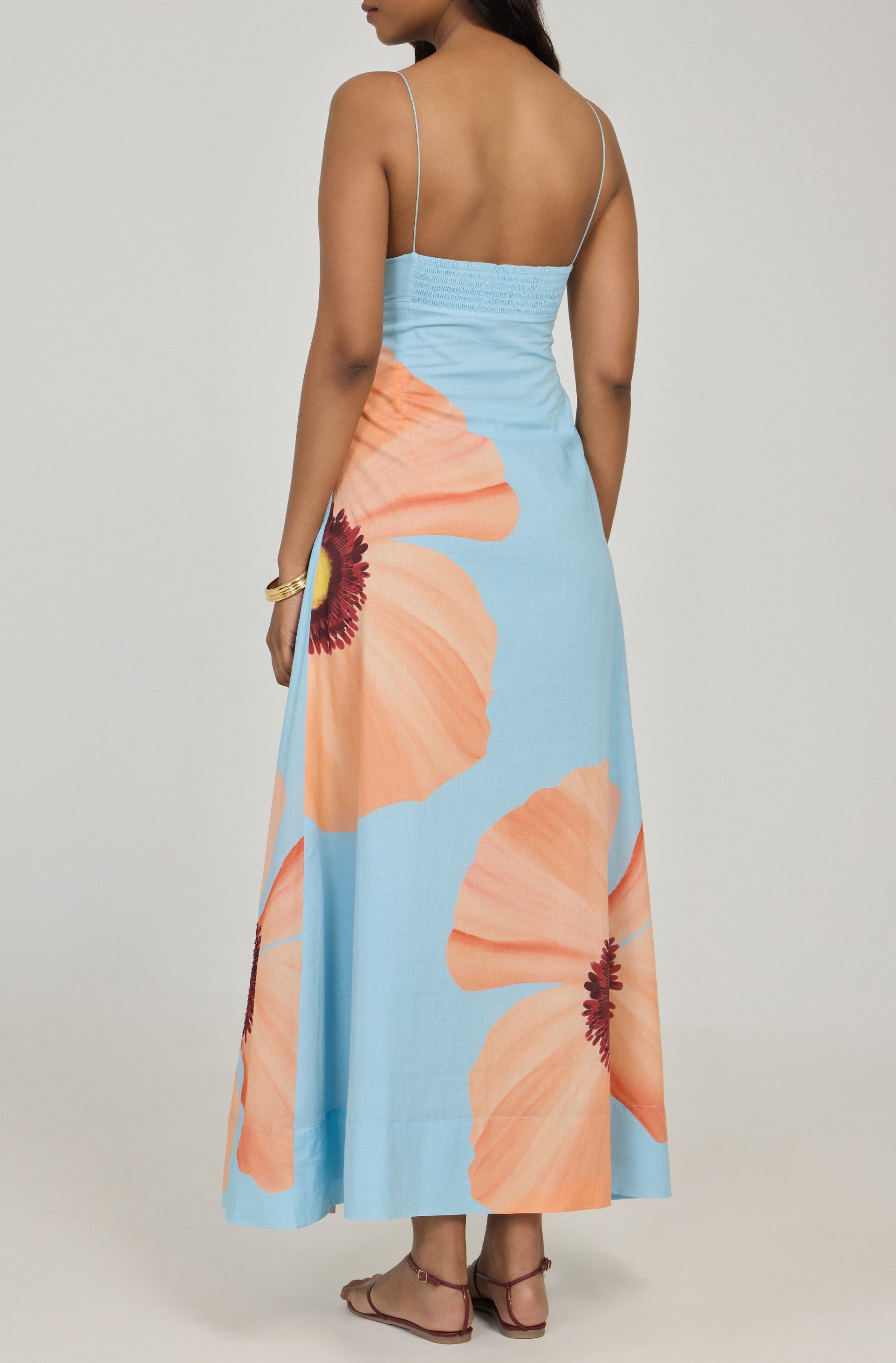 A woman stands facing away, wearing the Summer Away Alfama Maxi Dress with adjustable straps and peach floral prints, paired with flat sandals and a gold bracelet.