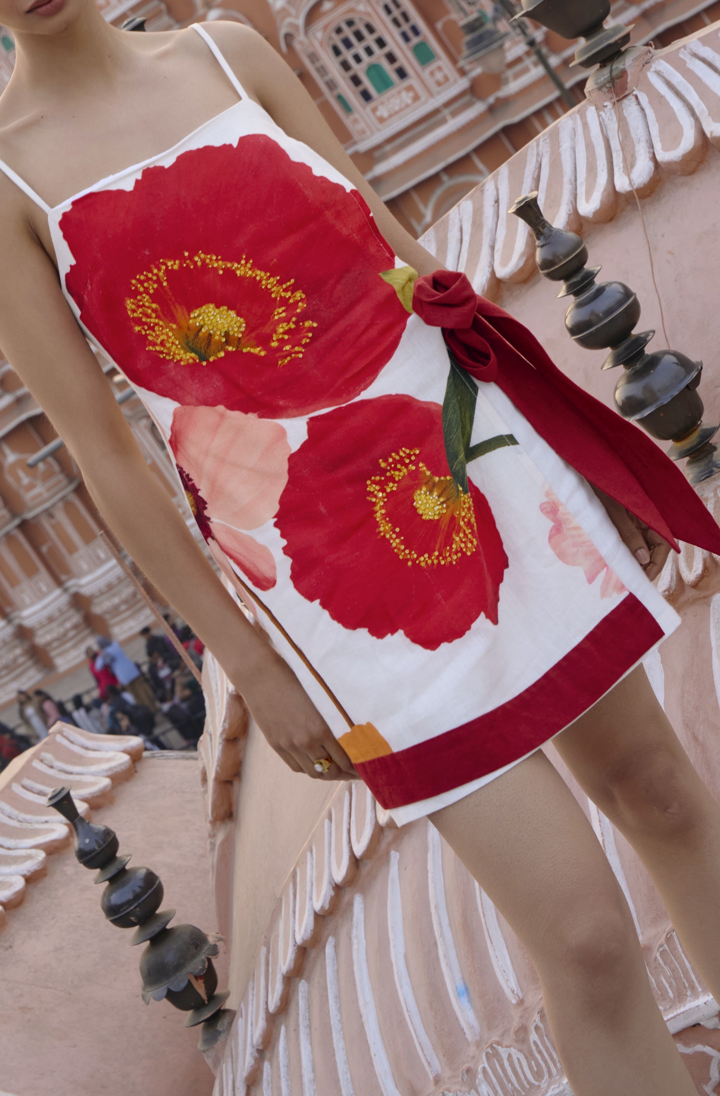 Wearing the Summer Away Anela Mini Dress, featuring a white faux wrap style with bold red floral prints, a woman stands on an ornate pink balcony with decorative railings and a historic building behind her.
