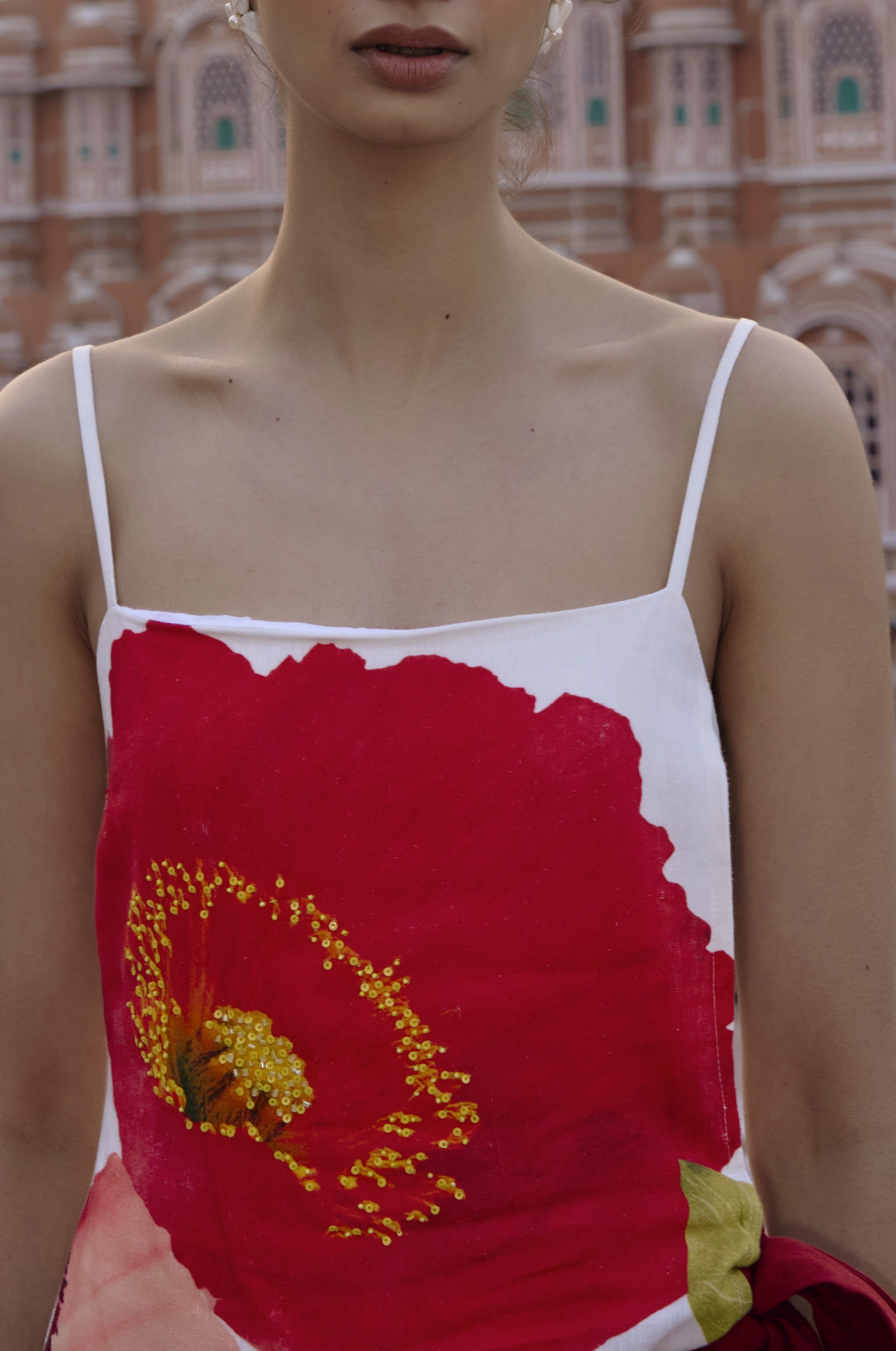 A woman in the Summer Away Anela Mini Dress—white linen with thin straps and a bold red flower print—stands outdoors, a blurred historic brown-and-white building in the background.