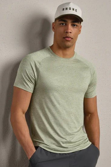 A man stands against a light-colored wall with his hands in his pockets, wearing a RHONE Atmosphere Tee in Dusty Sage Heather, dark gray pants, and a white RHONE baseball cap.