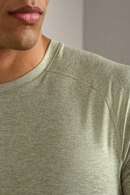 Close-up of a man wearing the RHONE Atmosphere Tee, a light olive-green, short-sleeve crew neck shirt crafted from lightweight performance fabric. Only the lower face, neck, shoulder, and upper chest are visible against a blurred background.